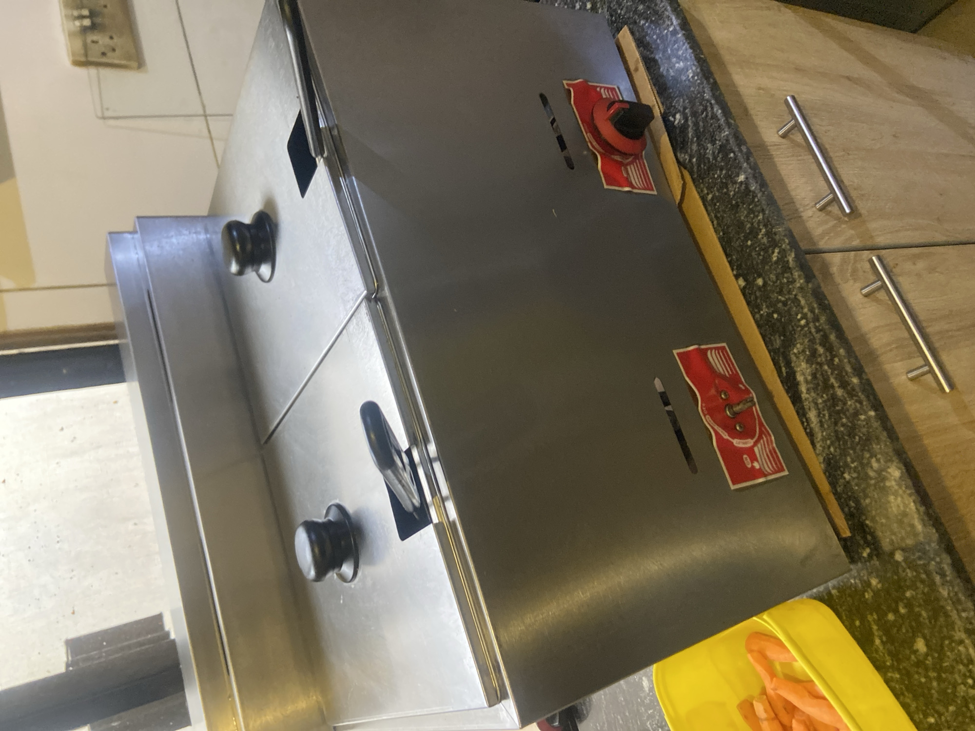 This image captures a kitchen or food preparation area in Howayi, Nigeria. The central focus is a dual-compartment commercial cooking appliance, likely a deep fryer, constructed from brushed stainless steel. The appliance features two rectangular top compartments, each with a flat, hinged lid equipped with a black, knob-style handle. A metal skimmer or basket handle with a black grip rests in a slot on the right lid. The dark grey front panel of the appliance displays two red rotary control knobs with black centers, positioned over red rectangular labels, one of which is partially obscured. Horizontal ventilation slits are visible above these controls. The appliance rests on a thin, light-colored wooden or cardboard strip, directly on a dark, speckled granite countertop.
To the left of the appliance, a bright yellow rectangular plastic container holds several irregularly cut, peeled orange vegetable pieces, consistent with carrots or sweet potatoes. Below the countertop, light-toned wooden cabinets are visible, fitted with horizontal silver bar handles. The countertop surface shows some scattered light-colored residue. In the background, an upper left area reveals a dark vertical frame and a light-colored, slightly textured wall or window. The upper right background features a light-tiled wall with visible grout lines and a cream-colored double electrical outlet. No people are present in the scene.