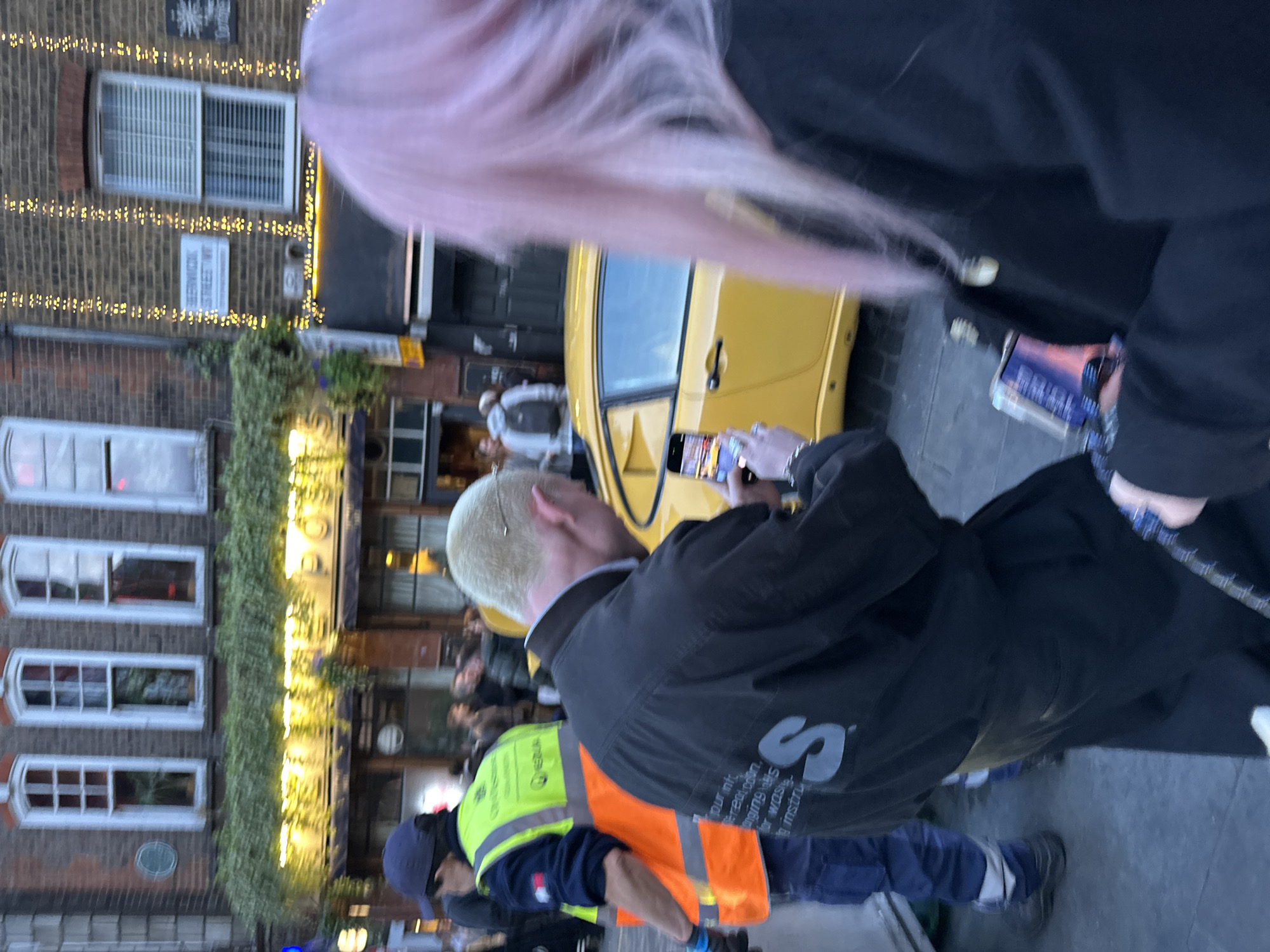 The image depicts a vibrant street scene in London during dusk or early evening, characterized by artificial lighting. The perspective is slightly tilted, capturing individuals and buildings along a bustling street.
In the foreground, a person with long, pink hair is partially visible from behind, wearing a dark top. Their smartphone is held in their hand, displaying a colorful image. Directly in front of them, another person with short, light blonde hair is seen from the back, wearing a dark jacket. A large grey 'S' logo is prominent on their back, accompanied by smaller white text that is partially legible, including what appears to be "terrean.com" and "mstr". This individual is actively holding up a smartphone, seemingly capturing a photograph or video.
Further down and to the bottom, two individuals are wearing high-visibility workwear. One wears a blue cap, blue work trousers, and a fluorescent yellow vest over a blue shirt, with text "MESQUA" and "CONS" partially visible on the vest. Another person wearing an orange high-visibility vest is partially obscured behind them.
The background features classic brick buildings, one adorned with strings of decorative warm-toned lights. Several windows with white frames and blinds are visible on this building. A clear white street sign is mounted on the brick wall, explicitly stating "HENRYCOCK STREET WC1," confirming the location in Central London. A bright yellow car is parked or moving in the midground. A vertical strip of green foliage climbs the side of another building. Blurred figures are visible further down the street, suggesting a moderate crowd. The scene conveys an active urban environment, possibly a social gathering or evening event.