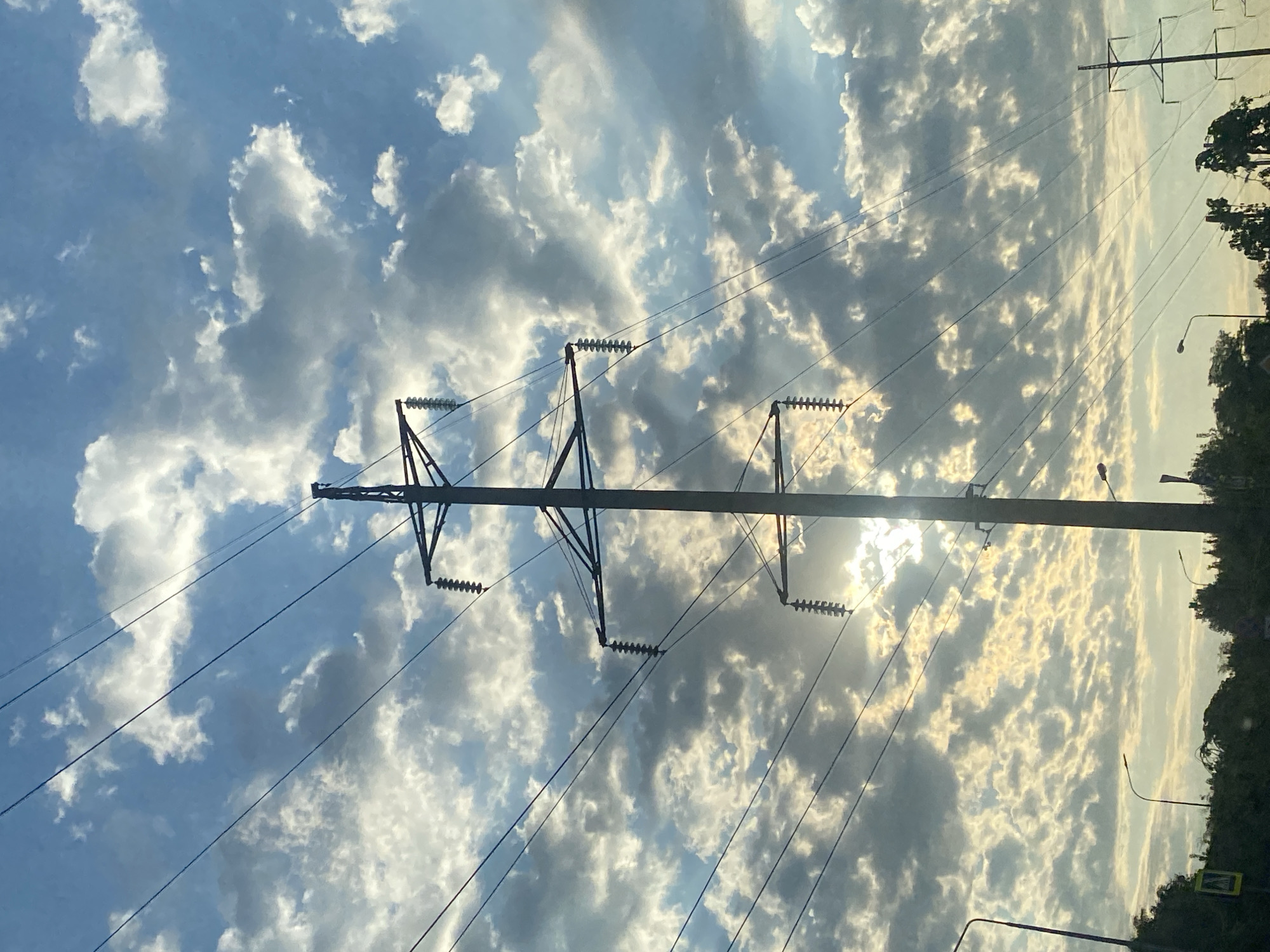 An outdoor, eye-level shot captures a tall, silhouetted power line tower against a bright, cloudy sky. The sun peeks through the dense cloud cover, casting a warm glow. Multiple power lines stretch diagonally across the frame, with insulators visible on the tower. In the foreground, the tops of dark green trees are visible, with streetlights and a pedestrian crossing sign also present, suggesting an urban or suburban setting.