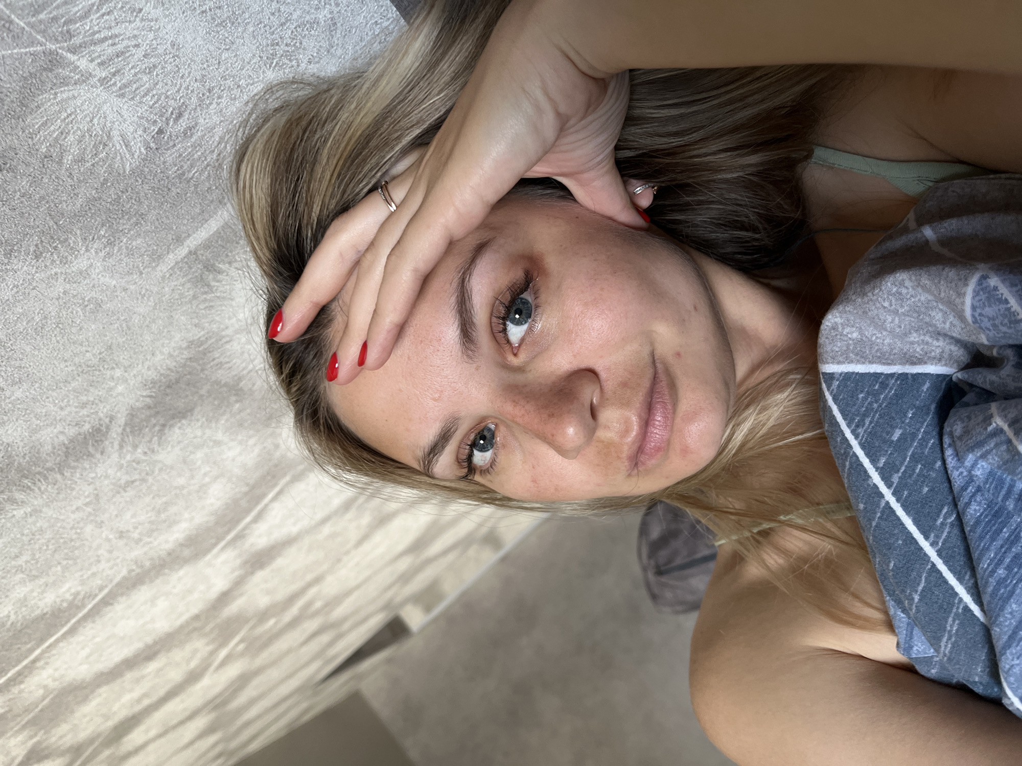 This is a selfie of a young woman lying in bed, taken at an angle from above. The woman has blonde hair and blue eyes. Her left hand is resting on her forehead, and she is wearing a ring on her left index finger. Her fingernails are painted red. She is wearing a light green bra or camisole, and is partially covered by a blue and white plaid blanket. The background appears to be a bedroom with textured walls or wallpaper and a beige floor. The lighting suggests it is daytime, possibly morning or evening. The woman's expression is relaxed and natural. There is no visible text in the image. The location context is Chaikovsky, Russia.