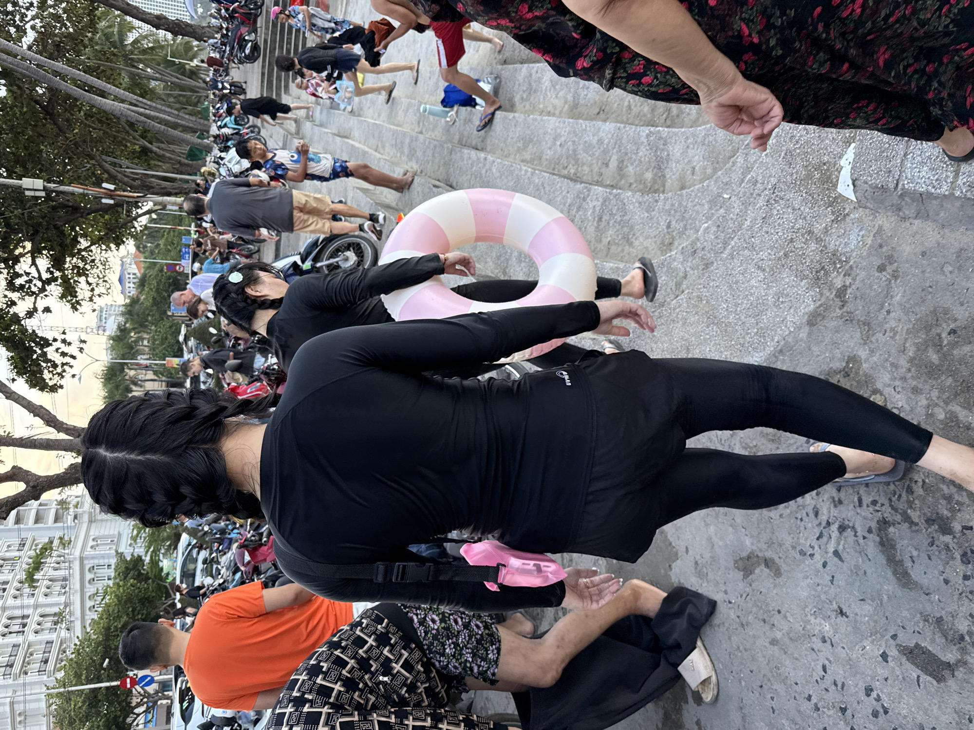 This image shows a group of people gathered near a waterfront area, likely a beach or promenade, in Nha Trang Ward, Vietnam. The foreground is dominated by the back of a woman with black braided hair wearing a black long-sleeved rash guard and black leggings. She is holding a pink waterproof phone pouch attached to her side by a black strap. To her left, another person is partially visible wearing a patterned long-sleeved shirt and floral shorts.
In the mid-ground, people are walking and standing on stone steps. A pink and white striped inflatable ring is being carried by one of the individuals. Several motorcycles are parked in the background, suggesting a busy urban or tourist area. Palm trees line the background, and a large, multi-story building with a white facade is visible in the upper left corner, typical of coastal resorts. The overall lighting suggests it is either late afternoon or early evening. The scene appears to be a public space where people are transitioning between activities, possibly heading to or from the beach, given the presence of the inflatable ring and the attire of some individuals.