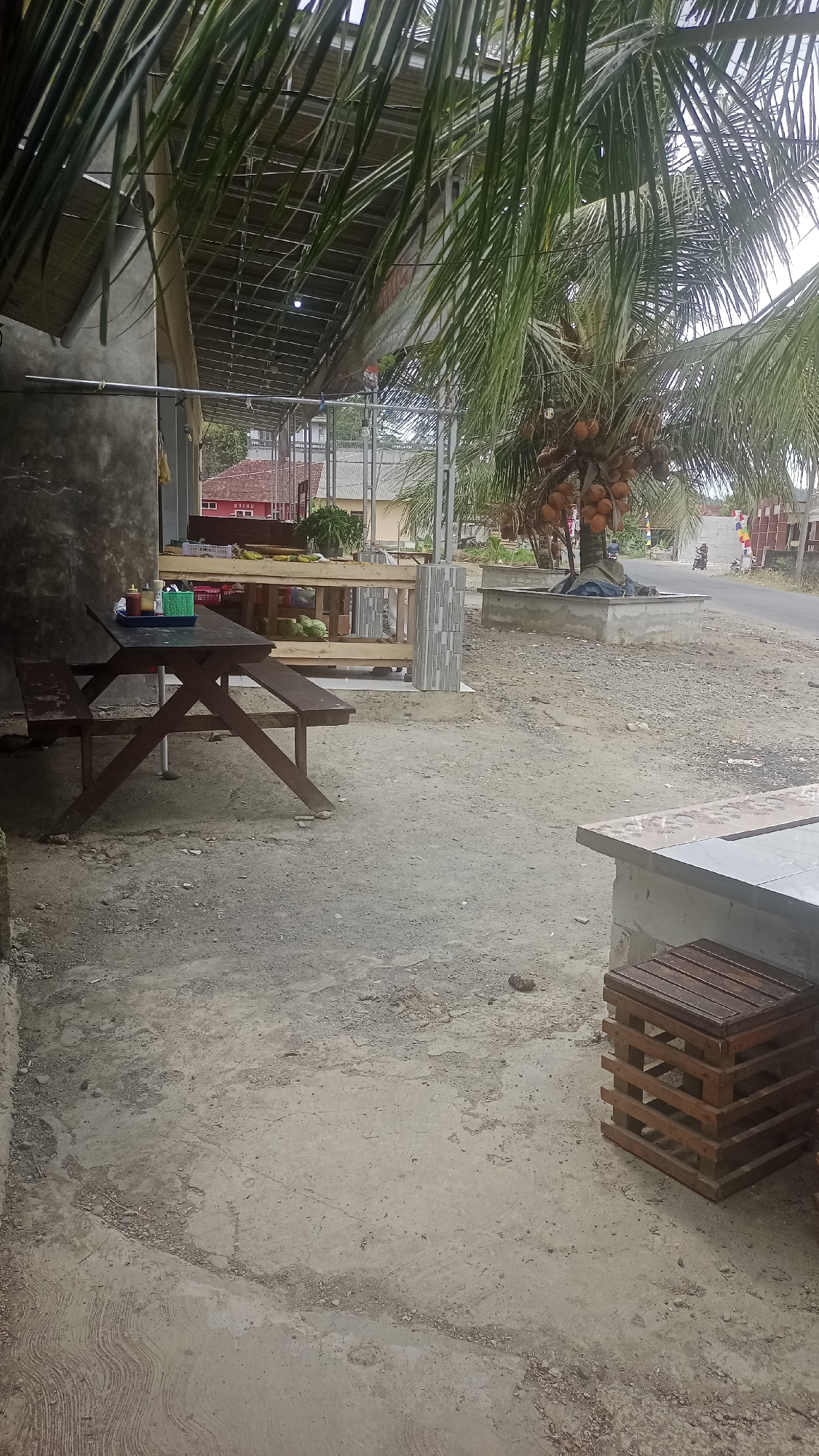 The image depicts an outdoor area adjacent to a building with a corrugated metal roof supported by metal poles. A picnic table with attached benches, made of dark wood, is positioned on a concrete ground. On the table, there are three small bottles of condiments and a green plastic container. In the background, a wooden stall displays what appear to be coconuts and other produce. Beyond the stall, there is a road and buildings. A coconut palm tree laden with fruit stands to the right of the stall. A wooden cubed object is visible in the foreground to the right. The ground is a mixture of concrete and dirt. The scene is likely a roadside eatery or market in Cibeber, Indonesia.