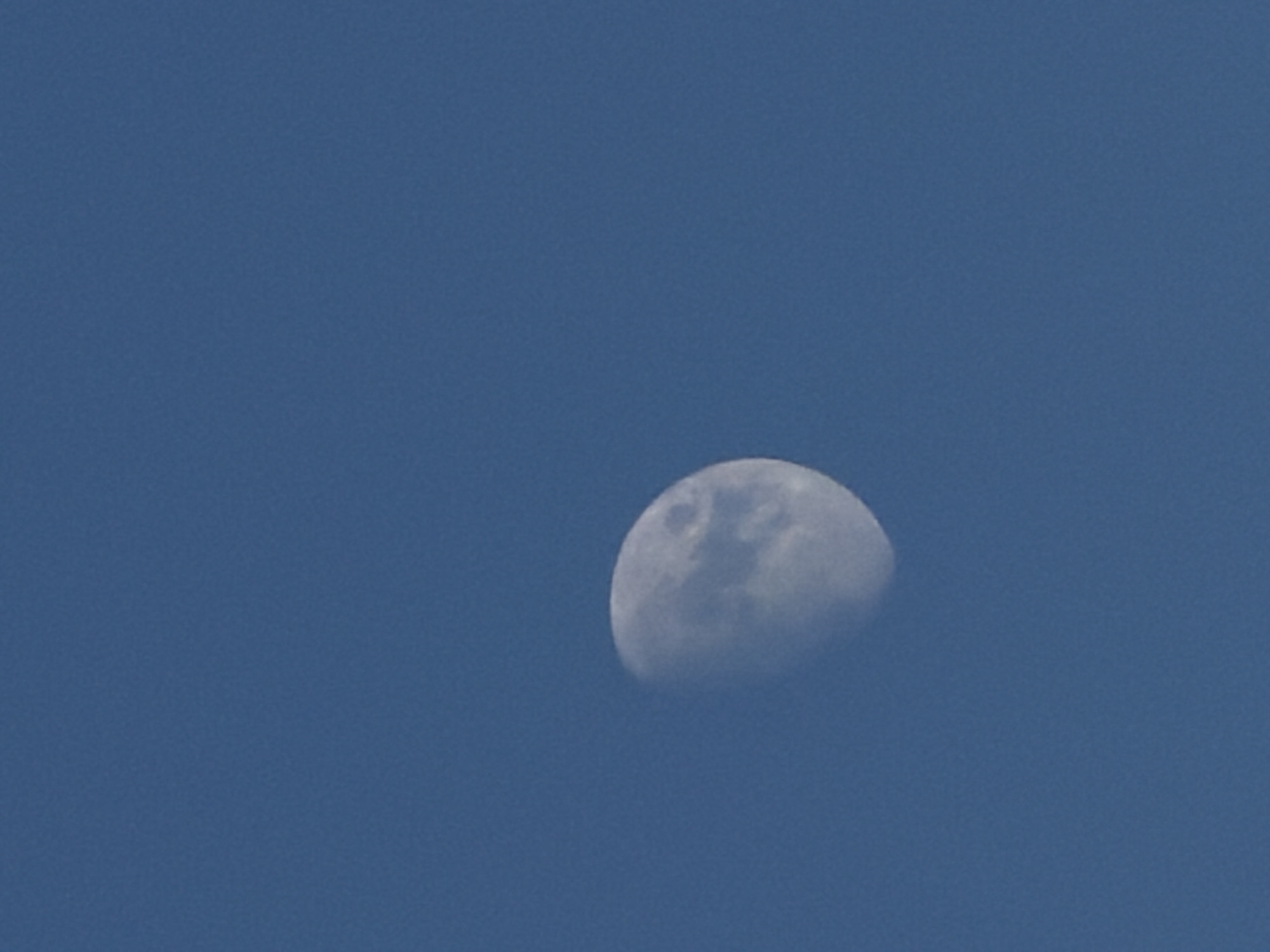 The image shows a portion of the moon against a clear blue sky. The moon is in its gibbous phase, with more than half of its surface illuminated. The illuminated part of the moon displays visible craters and surface features, suggesting it is daytime or twilight. The sky is a uniform shade of blue, with no clouds present. The image appears to be taken from a ground-level perspective, looking upwards. No people, text, or specific location cues are visible in the frame. The overall impression is of a clear sky with the moon prominently displayed.