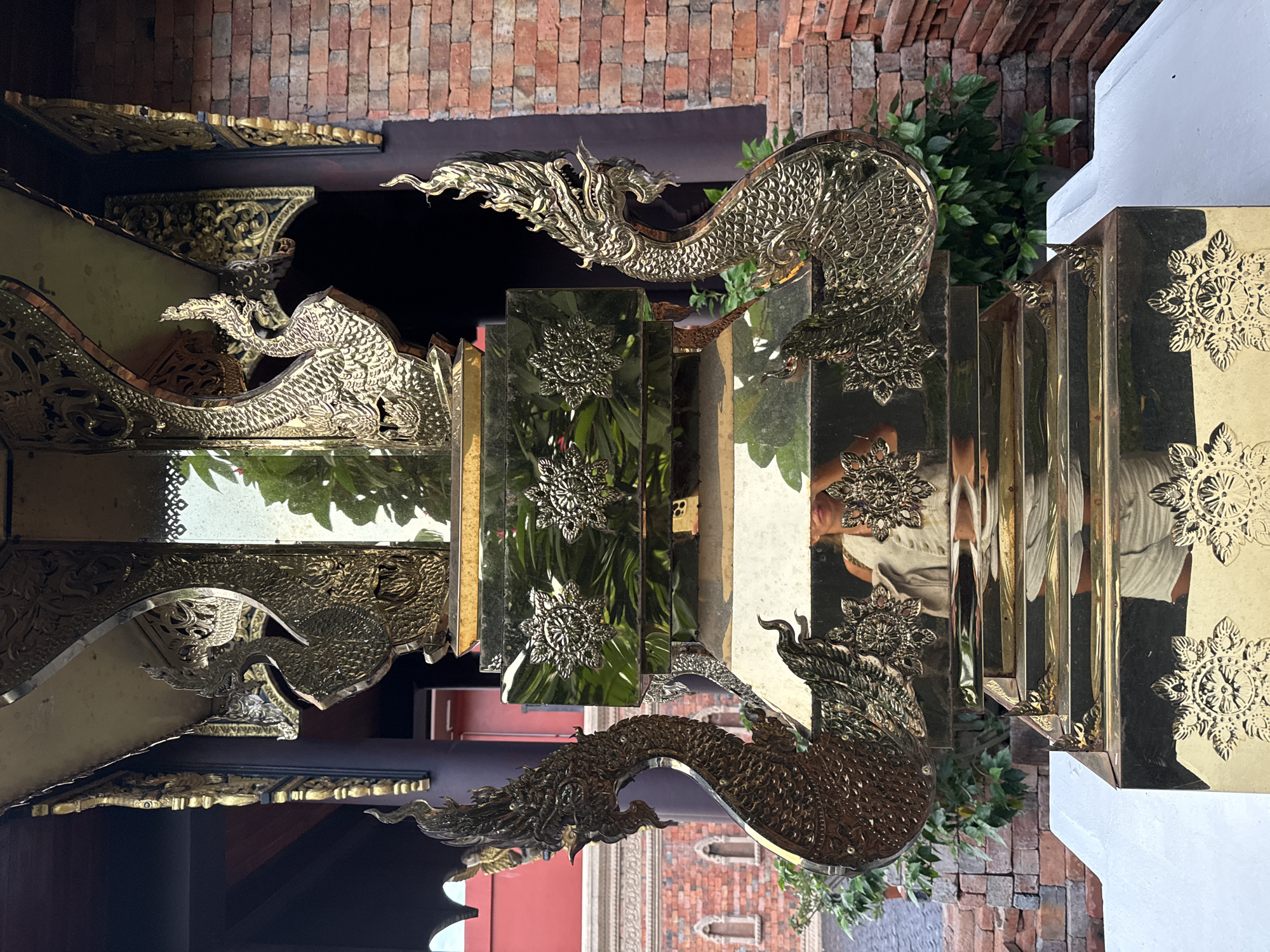 The image shows a highly ornate, gold-colored structure with intricate carvings of mythical creatures that appear to be Nagas, serpentine beings prominent in Buddhist and Hindu mythology. The structure is multifaceted and tiered, with a glossy, reflective surface that mirrors the surroundings and a person taking the photo. Decorative floral motifs are also visible. Behind the structure, a wall made of red brick and a dark, ornate roofline with golden embellishments are present. Lush green foliage is seen to the sides. The scene suggests an outdoor setting, possibly at a temple or a place of cultural significance in Ko Samui District, Thailand, given the architectural style and iconography. The photographer is reflected in the mirrored surfaces, wearing a light-colored top.