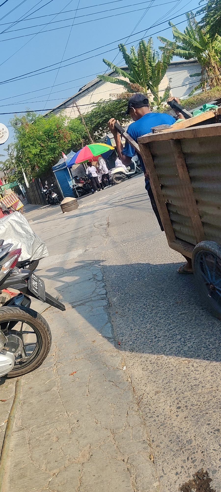 In Mekarmukti, Indonesia, on a sunlit day, a man wearing a blue t-shirt and a black baseball cap pushes a wooden cart with a corrugated metal side. The cart has a dark-colored wheel. In the background, there is a colorful umbrella covering a small stall. Several people are gathered under the umbrella, some of whom appear to be wearing white shirts. Motorcycles are parked nearby. Banana trees and other vegetation are visible, along with a building with a sloped roof. Power lines crisscross the sky. A portion of a motorcycle is in the foreground on the left, with its rear wheel and license plate visible. The ground is a mix of cracked concrete and asphalt.