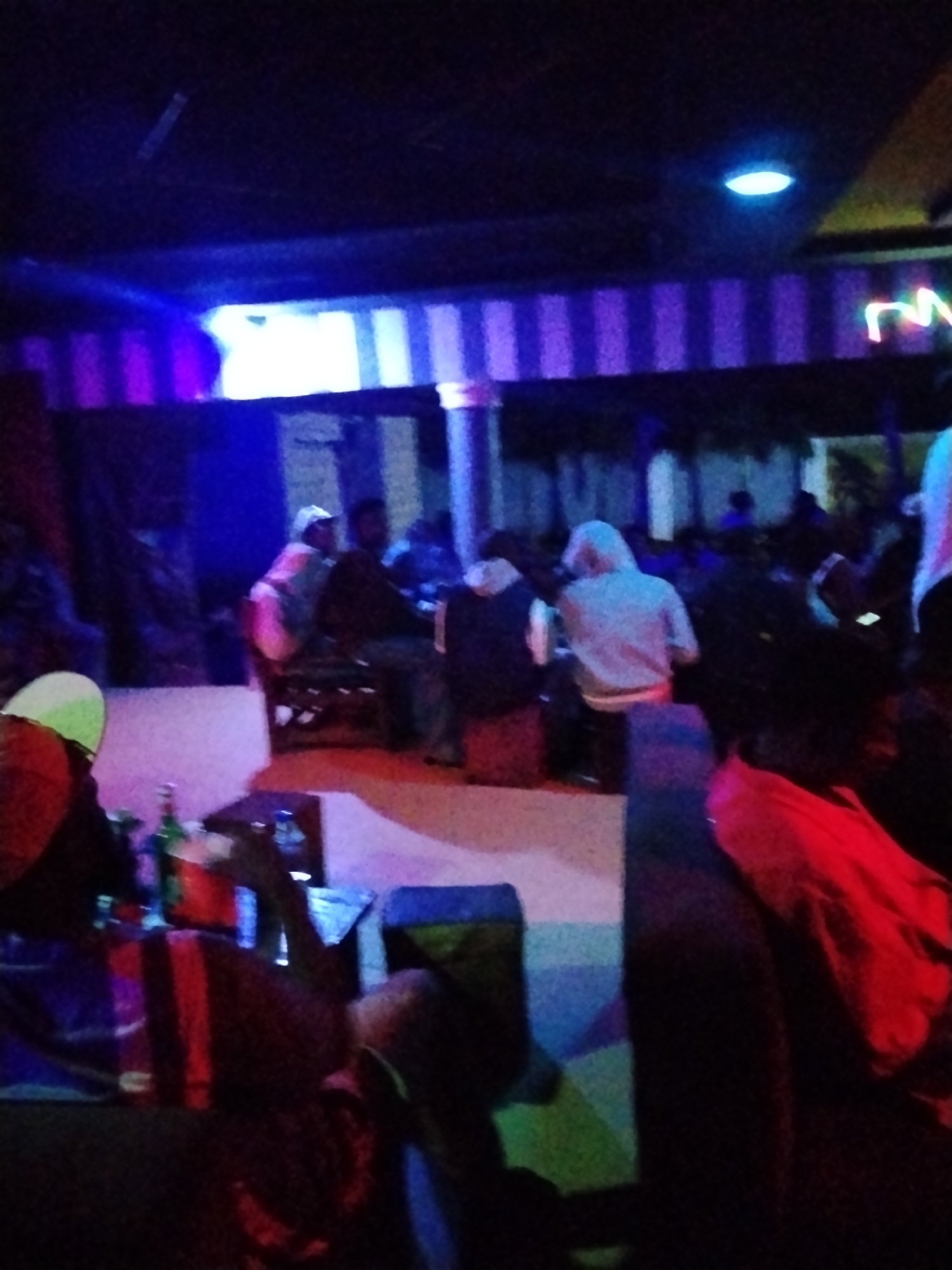The image shows a dimly lit interior of what appears to be a bar or lounge. There are several people present, seated and standing, some appearing to be engaged in conversation. The environment is characterized by neon lighting, with purple and blue hues dominating the scene, casting long shadows. In the foreground, a table with bottles and glasses is visible. The overall impression is that of a social gathering in an entertainment venue.