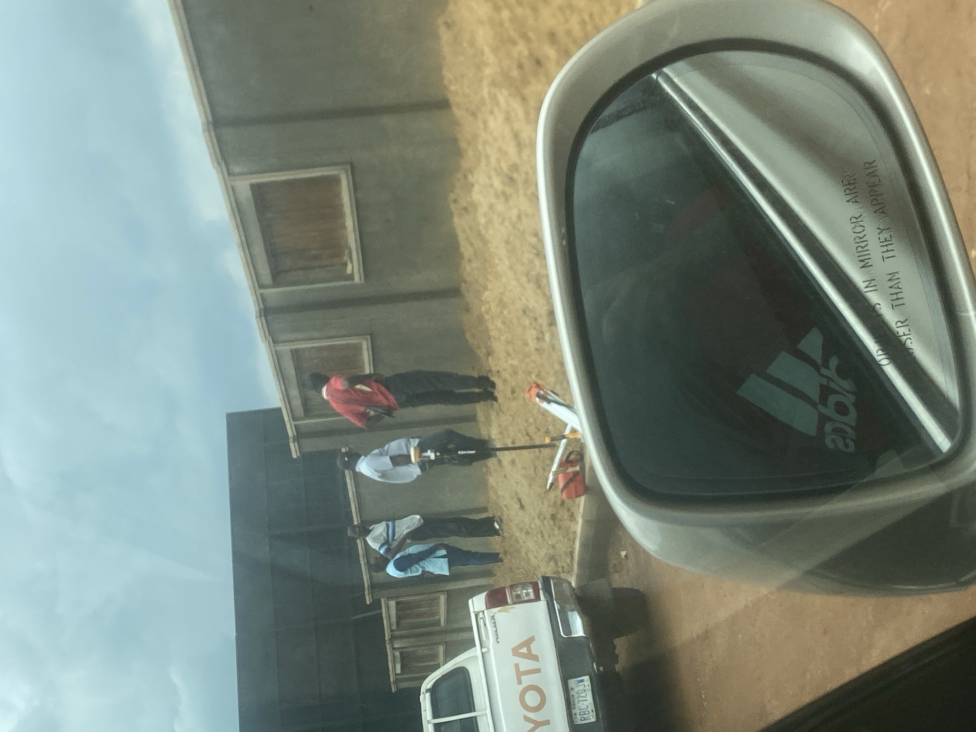 The image captures an outdoor scene, likely in Gosa, Nigeria, viewed from the perspective of a vehicle's side mirror. The metallic grey side mirror dominates the right side of the frame, prominently reflecting an Adidas logo and the partially visible text "OBJECTS IN MIRROR ARE CLOSER THAN THEY APPEAR."
Beyond the mirror, four men are standing on a dry, dusty, or gravelly ground surface. One man in a red shirt stands to the left. Central to the group is a man in a white shirt operating or observing a tripod-mounted piece of surveying equipment, possibly a total station or GPS receiver. A prism pole with red and white markings stands upright nearby. Two other men, wearing a striped light blue shirt and a solid light blue shirt, are positioned further to the right.
A white Toyota Hilux pickup truck is parked facing away from the viewer, visible towards the bottom right. Its tailgate clearly displays the "TOYOTA" and "HILUX" branding, along with a Nigerian license plate reading "RBC-720-JW". In the background, a light-colored building with multiple rectangular window openings, some appearing boarded or covered, stretches across the scene. The sky above is bright and partly cloudy, indicating daytime. The activity suggests the men are involved in land surveying or site preparation work.
