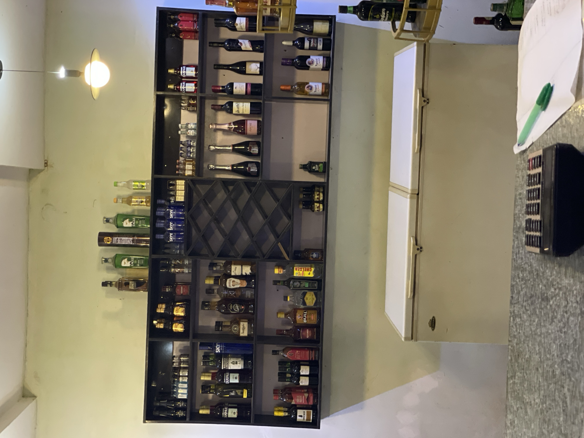 The image shows a well-stocked bar area in what appears to be a commercial establishment, likely in Akwanga, Nigeria. A large black shelving unit is filled with a variety of alcoholic beverages, including wines, spirits, and liquors. Brands such as SKY vodka, Campari, and others are visible. Next to the shelves is a light-colored freezer. On the right, a countertop is visible, with items such as paper and a black object (possibly a cash register or other electronic device) resting on it. A light fixture is positioned on the left, illuminating the scene. The walls are painted in a light color.