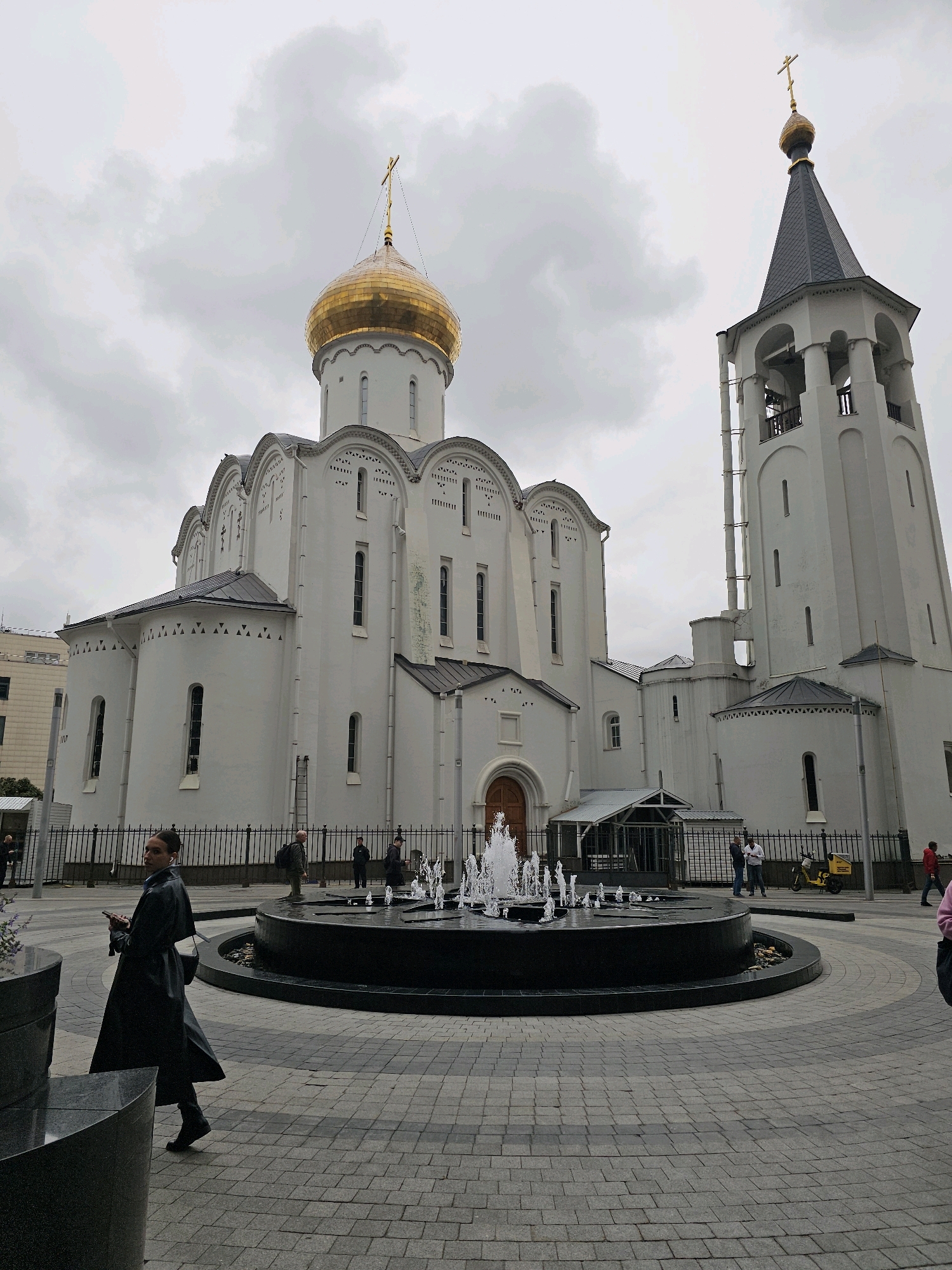 The image shows the Church of the Resurrection of Christ, located in Moscow, Russia. The church is a white building with a golden dome and a tall bell tower. In front of the church is a large fountain with water spraying upwards. A person in a black coat walks past the fountain, looking at their phone. Several other people are visible in the background, as well as a black metal fence and some trees. The sky is overcast.