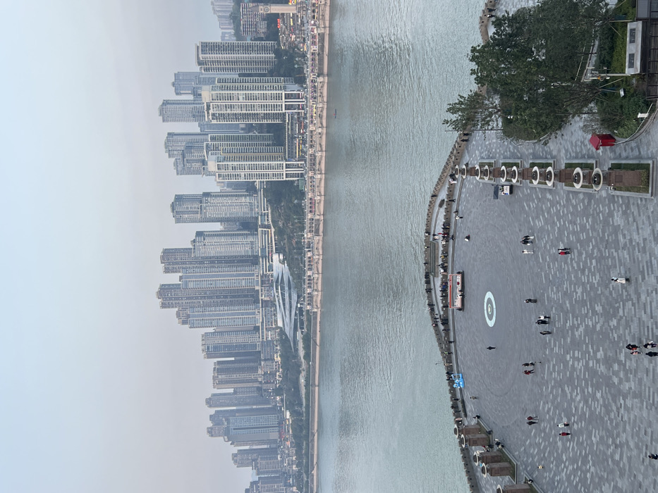 The image presents an elevated panoramic view of a riverfront area in Yuzhong District, Chongqing, China, under a uniformly light grey, overcast sky.
In the foreground, a large, irregularly shaped public plaza features a paved surface composed of grey, rectangular interlocking tiles. A distinct circular motif with a central blue disc is embedded in the pavement near the plaza's center. Numerous individuals, estimated between 75 and 100, are scattered across the plaza. A prominent gathering of approximately 30-40 people is observed along the curved, crenellated stone wall that forms the plaza's edge overlooking the river, with most individuals facing the water. Other individuals are seen walking across the plaza in various directions, some in small groups. Near the river-facing edge, a white, rectangular kiosk or temporary booth is visible, alongside a larger blue inflatable structure. Along the right side of the plaza, a series of decorative, column-like structures with circular flat tops are symmetrically spaced within landscaped planter boxes, accompanied by small trees and shrubs. A red utility box is visible within the greenery on the far right. A small utility vehicle or cart is positioned on the plaza's right side.
The midground is dominated by a wide, calm body of water, likely the Yangtze River, appearing grey-green under the subdued light. A single small, dark-hulled boat is visible on the water, moving away from the viewer. Along the opposite bank, a multi-arched bridge or waterfront promenade structure extends horizontally across the scene.
The background consists of a dense urban cityscape ascending a verdant hillside. Numerous high-rise buildings and skyscrapers, primarily constructed from light-colored concrete and glass, dominate the skyline. These structures appear to be a mix of residential and commercial properties. A distinctive, large modern building with multiple curved tiers and a white façade is visible towards the left-center, likely a prominent civic or cultural landmark. On the far right, a building with a prominent clock tower is discernible. The urban development extends deeply into the distant, hazy horizon.