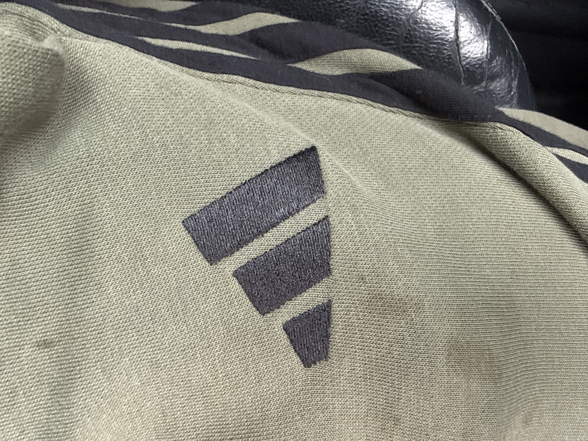 The image shows a close-up of a light olive-green Adidas garment with the brand's three-stripe logo in a darker grey or black. The logo consists of three parallel bars angled upwards and to the right, decreasing in size from top to bottom. The garment appears to be made of a knit fabric with a textured surface. A section of what appears to be a black leather or synthetic material is visible in the upper right corner, potentially part of a seat or another article of clothing.