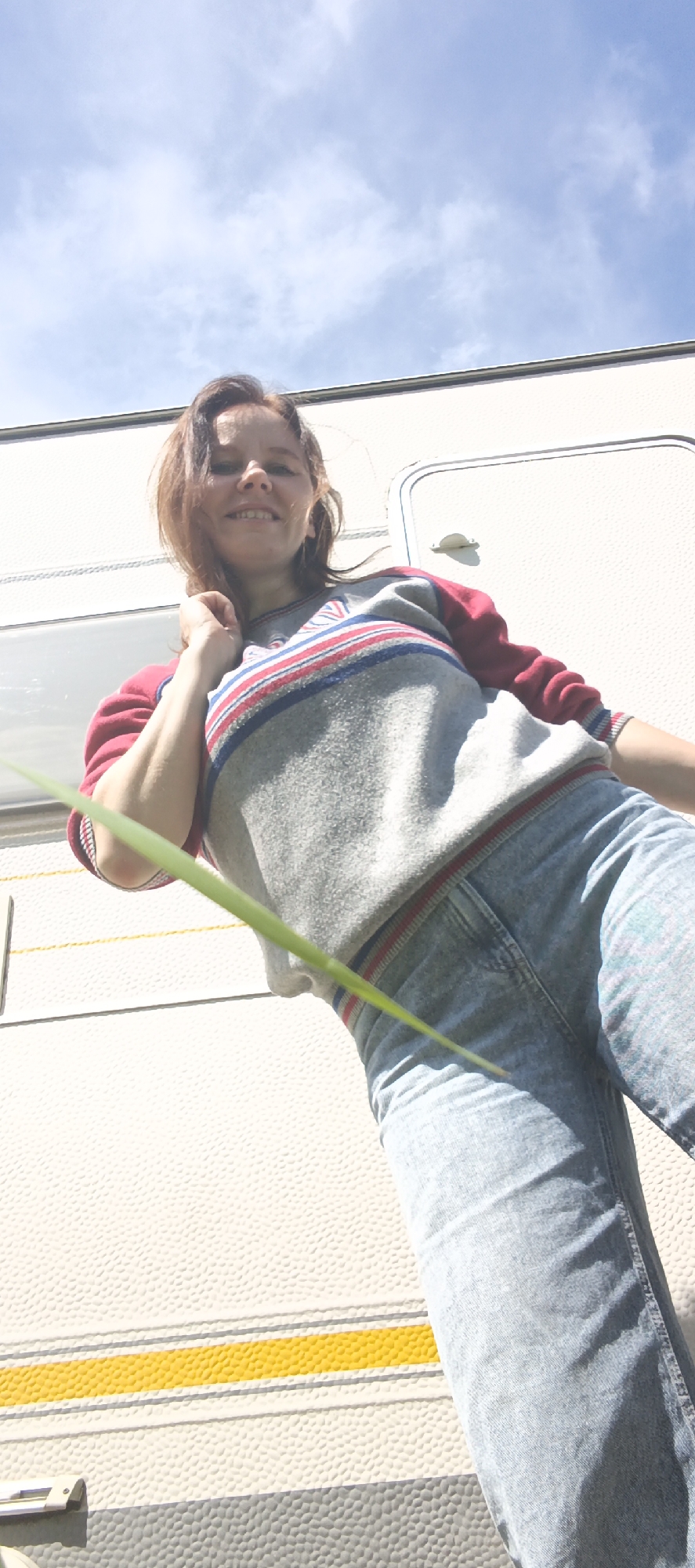 A low-angle, full-body shot depicts a young woman posing outdoors. She is wearing a grey sweatshirt with red and blue stripes on the sleeves and collar, and light blue denim jeans. The woman has medium-length brown hair and is smiling at the camera. Her left arm is bent, and her hand is touching her neck. She is standing in front of a white recreational vehicle (RV) or caravan, which has a textured surface and horizontal stripes, including a prominent yellow stripe. A green blade of grass or leaf is positioned in the foreground, partially obscuring the woman's body. The background shows a bright blue sky with scattered white clouds. The scene appears to be in a park or campsite setting. The image does not provide specific indicators of its location within Chaikovsky, Russia, beyond it being an outdoor environment.