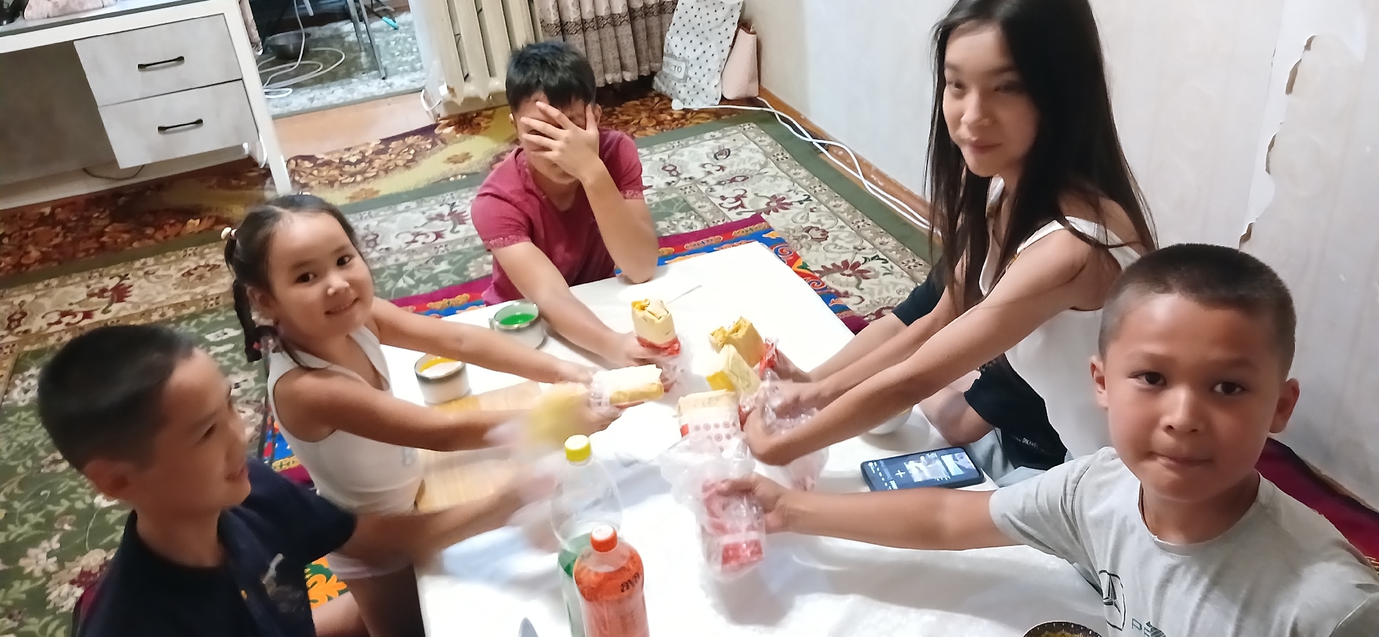 The image shows five individuals, four children and one young adult, gathered around a table in what appears to be a residential setting. The group is engaged in an activity involving food items, likely snacks or treats wrapped in plastic. The room is carpeted with a traditional patterned rug, and a white table is the central focus. Behind the group, a desk with drawers and a white radiator are visible, suggesting an indoor domestic environment. The lighting is ambient, creating a warm and casual atmosphere. The individuals are of varying ages, with the youngest appearing to be a toddler, and the oldest a young woman. The younger children are actively interacting with the food items, while the older individuals observe or participate in a more subdued manner. One child is covering their face with their hands, possibly in a playful gesture. A smartphone is also visible on the table, displaying an image or video. The scene suggests a family gathering or a casual get-together among friends. There is no specific indication of the location within Bishkek City, Kyrgyzstan, beyond it being an indoor residential space.
