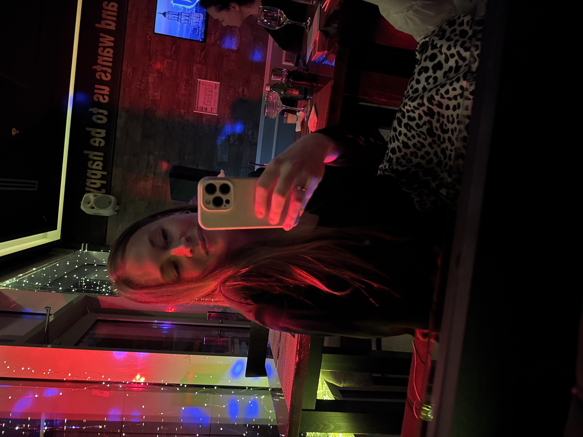 A selfie is taken inside a bar in Budva, Montenegro. A young woman holds a smartphone, her face illuminated by the screen. Red and blue string lights are visible through a window behind her. Another person is sitting at the bar with a drink in the background. A wall with writing on it and a television are also visible.