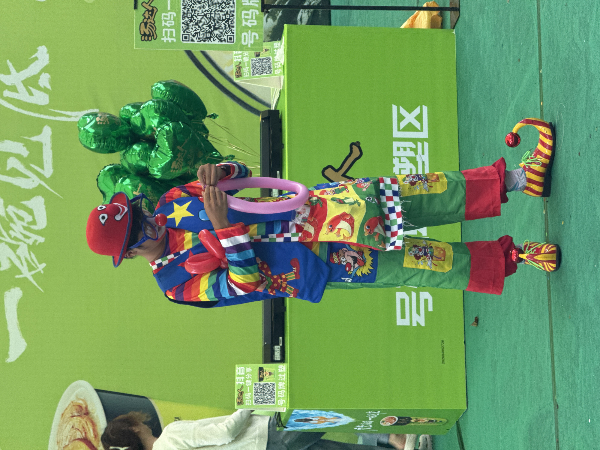 The scene is a public event in Nan'an District, China, featuring a clown in full costume. The clown is wearing a red hat, blue sunglasses, and a rainbow-striped jacket. They are holding a pink balloon in their hands. The clown's pants are green and red with cartoon patches. The shoes are yellow and red striped with curled toes. The clown is standing next to a green rectangular structure with Chinese writing on it. A bunch of green balloons are attached to the side. At the bottom left, there is a partial view of a woman's head and shoulder in a white top.