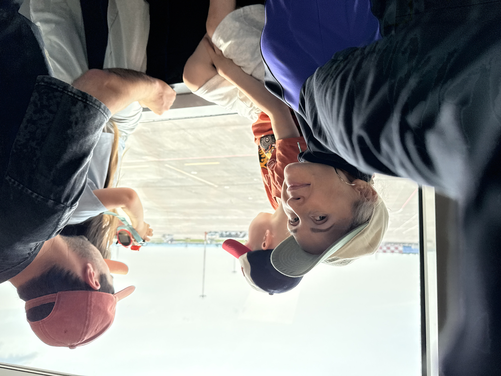 In the city of Bol'shie Kabany, Russia, a group of people, including at least three children and two adults, are visible looking out a large window, likely at an airport tarmac. A woman in a beige cap and a black jacket with a purple undershirt is in the foreground, her face turned towards the camera, smiling. Next to her, a child in an orange t-shirt and white shorts looks out the window, wearing a red and blue baseball cap. To their right, a man in a red baseball cap and a dark, textured t-shirt is holding a red toy camera, pointing it towards the window. A younger child, partially obscured, is also visible in front of the man. They are seated, with the window offering a view of an overcast sky, runway lights, and some distant airport buildings. The overall scene suggests a family waiting for a flight.