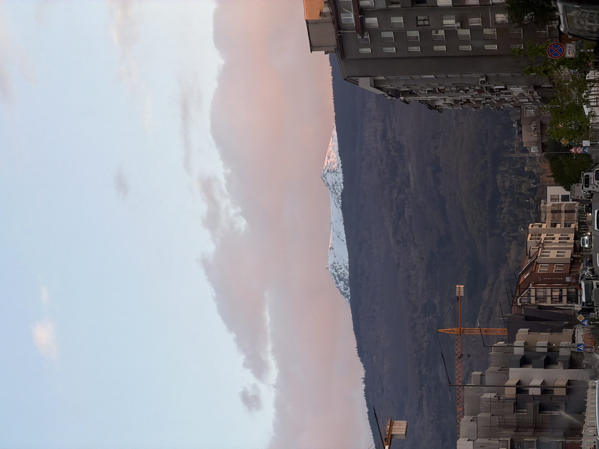 The image, correctly oriented, depicts a dynamic urban landscape in Sofia, Bulgaria, juxtaposed against a prominent mountain range under a colorful sky. The time of day appears to be either sunrise or sunset, indicated by the soft, warm pink and orange hues tinting the clouds and the distant snow-capped mountain peak.
In the midground and foreground, on the right side of the frame, a dense cluster of multi-story buildings lines what appears to be a city street. Some buildings exhibit typical Eastern European architecture with numerous windows and balconies, while others are clearly under construction, featuring scaffolding and an active yellow tower crane reaching into the sky. Below these buildings, parts of a street are visible, with standard European road signs including a "No Stopping" symbol, a "No Entry" symbol, a "30" speed limit sign, and a pedestrian crossing sign. Vehicles, including a white van, are partially visible on the street. On one building, faint Cyrillic text, possibly "ГРАД ОТЛОВ" (GRAD OTLOV), can be discerned.
Dominating the background is a vast mountain range. Its lower and mid-slopes are densely covered with dark green forest, conveying a sense of untouched nature. A distinct peak in the distance is prominently snow-capped, its white surface brightly illuminated by the low sun, creating a striking contrast with the shadowed forested areas. The sky above is partly cloudy, transitioning from a light blue at the top to warmer pink and orange tones closer to the horizon, reflecting the atmospheric conditions of dawn or dusk. No people are explicitly visible in the frame.