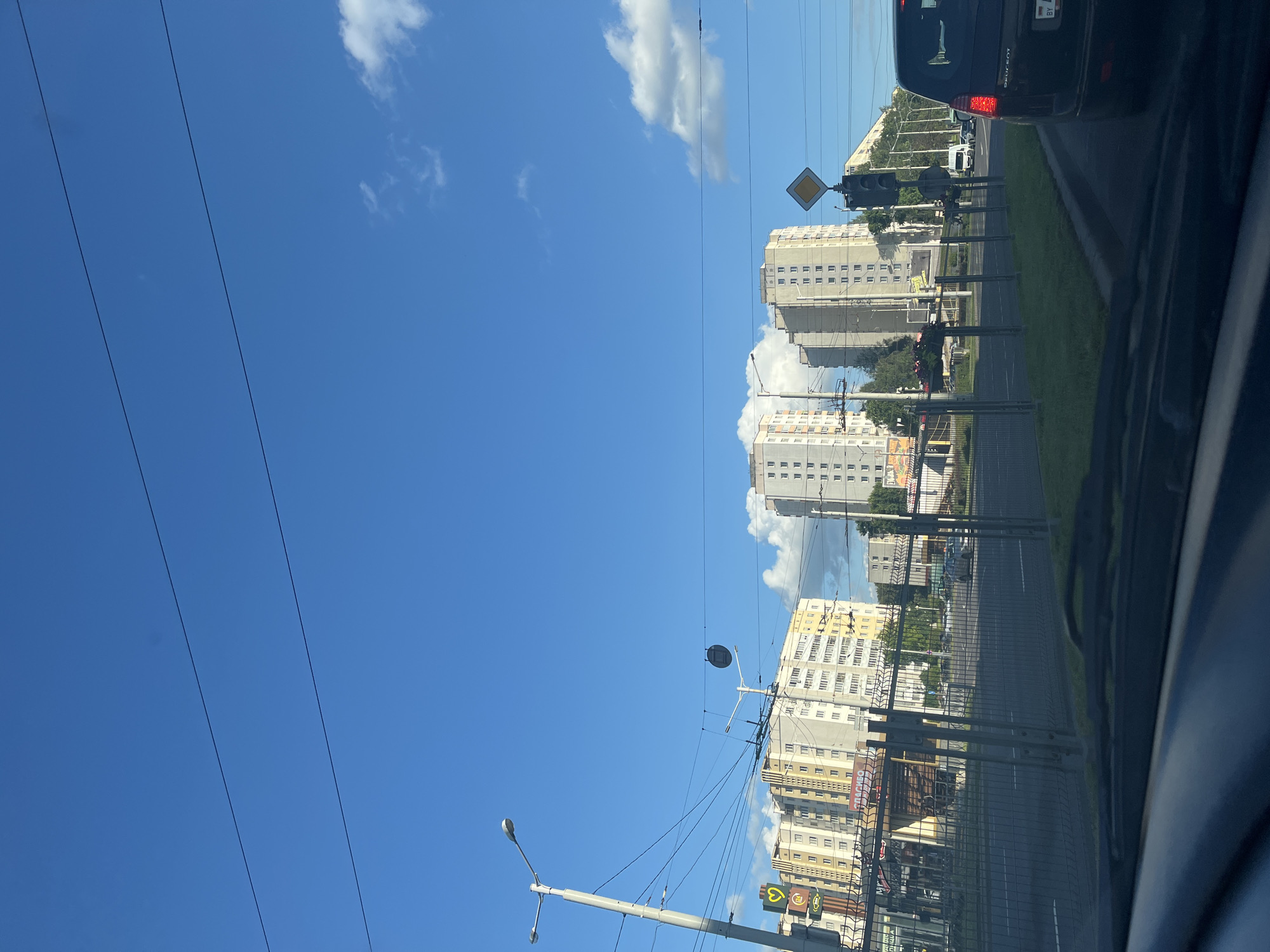 This is a landscape, eye-level, outdoor shot taken from a vehicle, looking forward through the windshield at a city street scene in Minsk, Belarus. The sky is a clear, bright blue with a few scattered white clouds. Three tall, light-colored apartment buildings dominate the midground, with several rows of windows visible on each. In the foreground, a metal fence runs horizontally across the frame. Beyond the fence, a paved road is visible with cars driving on it. A black Peugeot SUV is in the right lane, its taillights illuminated. Traffic lights and street lamps are also visible along the street. A yellow diamond-shaped road sign is mounted on a traffic signal pole. The scene appears to be a typical urban environment, likely a residential area with commercial establishments bordering the street.