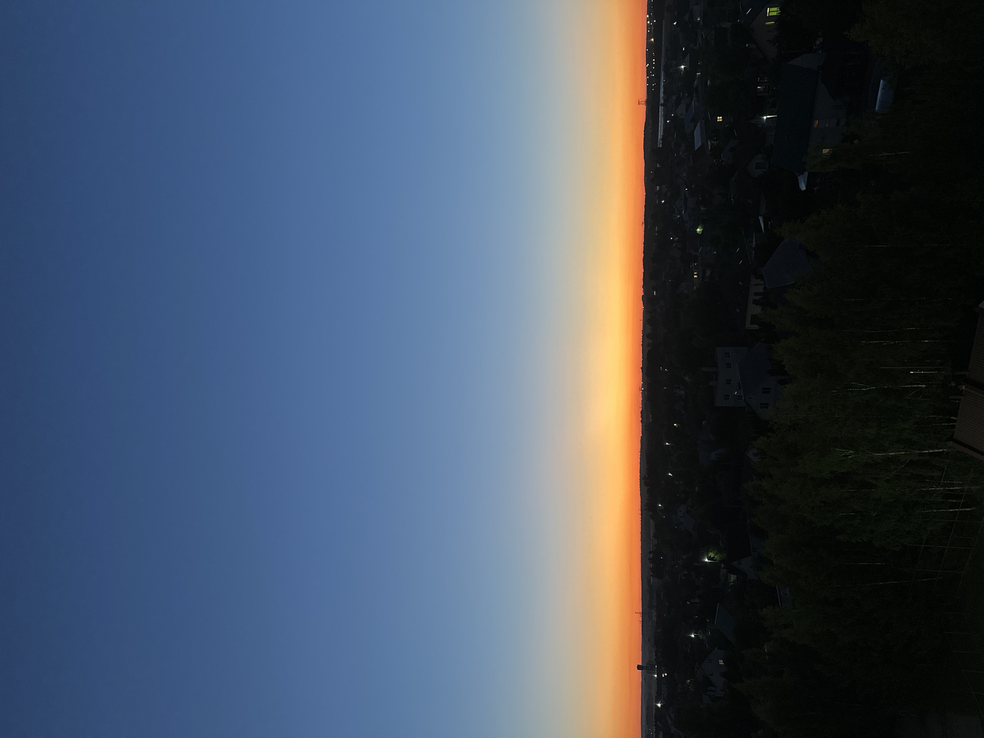 The image depicts a twilight scene over a suburban area in the vicinity of the village of Chomyśliski, Ščomiślicki sel’ski sovet, Belarus. The sky transitions from a deep blue at the top to a vibrant orange glow along the horizon, indicating the period just after sunset or before sunrise. Below the sky, a dark silhouette of a residential area is visible, characterized by numerous houses with scattered illuminated windows. A line of trees forms a dense, dark band in the foreground, obscuring the lower parts of the houses and suggesting they are located in a valley or at a lower elevation than the viewpoint. In the distance, faint outlines of industrial structures or tall towers are discernible against the bright horizon. The overall impression is of a quiet, rural or suburban landscape at dusk. There are no people or animals visible in the frame.