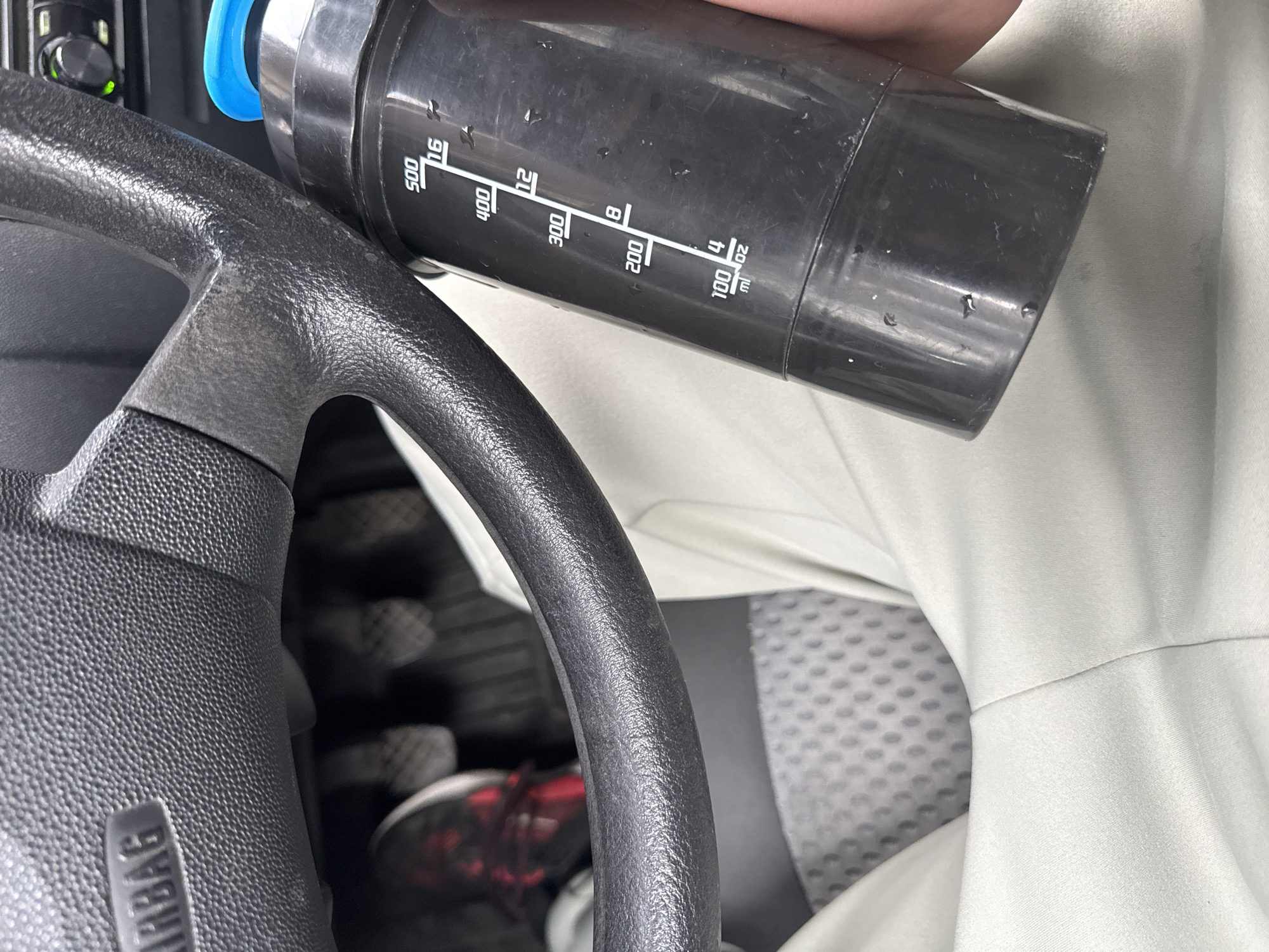 The image depicts the interior of a car, specifically the driver's side. A black steering wheel occupies the left foreground with an "AIRBAG" inscription visible. A blue-lidded black shaker bottle with measurement markings in milliliters (mL) and ounces (oz) sits near the steering wheel and the driver's leg clad in light-colored pants. The bottle's measurement markings are visible, suggesting it is partially filled. In the background, there's a view of the car's pedals and a glimpse of the car seat cover. The location cannot be determined from the image.