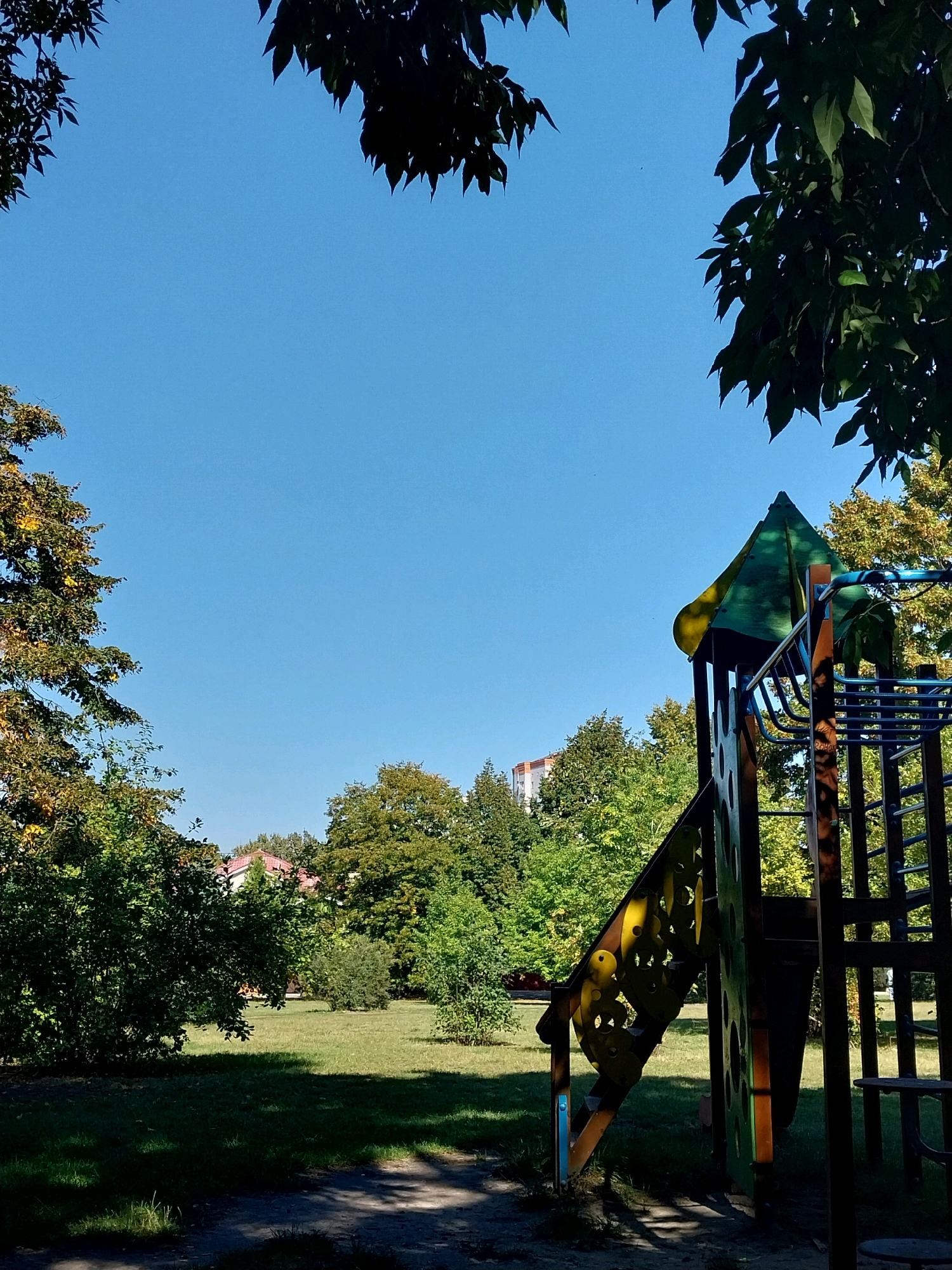 The image shows a playground with a bright blue sky above. In the foreground, to the right, is part of a colorful playground structure, featuring a green and yellow roof, a slide with yellow cutouts, and a blue railing. To the left and in the background, lush green trees with some autumnal yellowing foliage are visible. In the distance, a building with a red roof can be seen peeking through the trees. The ground is covered in grass, with shadows cast from the trees and playground equipment, suggesting it is daytime. The image is captured from a low angle, looking upwards. The scene appears to be in a park or residential area.