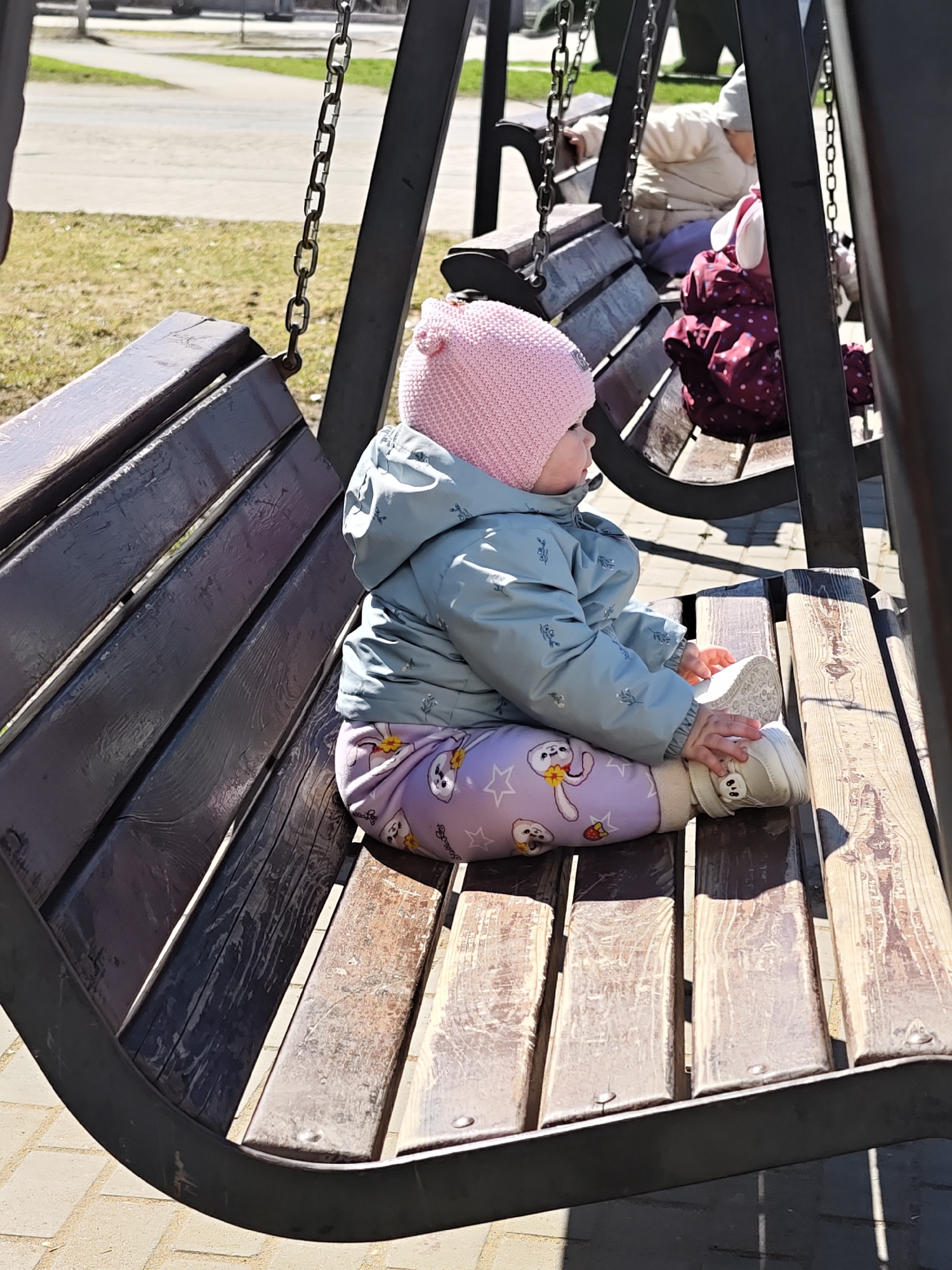 A young child wearing a pink knit hat and blue jacket sits on a wooden swing bench. The child is wearing patterned leggings and shoes, and is holding a shoe in their hands. Another swing bench with a child is visible in the background, and the scene is outdoors on a sunny day, likely in a park. The location is Chaikovsky, Russia.