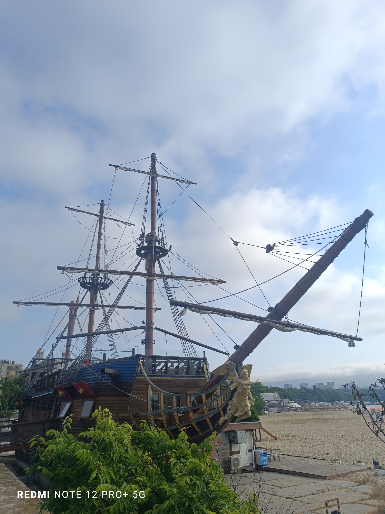 The media file presents a striking image of a large, multi-masted wooden ship, designed in the style of a historic galleon or pirate vessel, positioned on land next to a sandy beach. The ship is elaborately detailed, featuring tall masts with rigging and furled sails, wooden planking, and a distinct blue-colored roof section over its stern cabins. A prominent golden figurehead, depicting a winged female figure, is visible on the bow.
The setting appears to be a coastal area, likely a resort or tourist destination. The ship is situated directly next to a wide, clear sandy beach. Across the beach, in the background, a town or city with numerous buildings, including some modern structures, stretches along the coastline and up a distant hill. Lush green vegetation partially obscures the foreground and the base of the ship.
The sky is bright with scattered white and light gray clouds, indicating a partly cloudy daytime with soft, diffused light. There are no people visible in the image, suggesting a calm or early hour. The ship itself appears to be a stationary structure, possibly functioning as a themed restaurant or attraction, rather than a seafaring vessel. A small white building with an air conditioning unit and a freezer unit (bearing the text "ICE CREAM") is visible near the ship's bow, supporting the idea of a commercial establishment.
A watermark displaying "REDMI NOTE 12 PRO+ 5G" is present in the bottom-left corner of the original image. The location is consistent with Varna, Bulgaria, suggesting this is a themed establishment on the Black Sea coast.