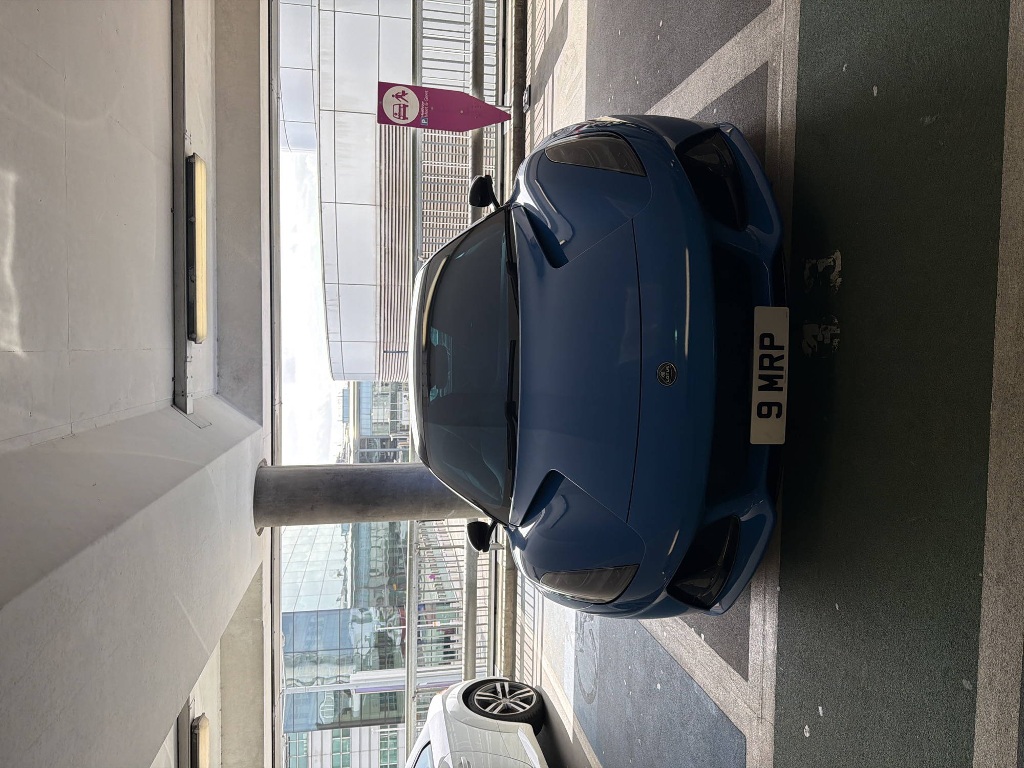 An image, rotated ninety degrees counter-clockwise, depicts a blue Lotus sports car parked in an indoor multi-storey car park, likely in London, United Kingdom. The car is positioned centrally, facing away from the viewer, with its front end visible. The license plate "9 MRP" is clearly displayed on the front bumper, below the Lotus emblem.
The setting appears to be a "Meet & Greet" parking area, indicated by a prominent pink, arrow-shaped sign hanging above the car. This sign features icons of a person and a car, along with the text "P Meet & Greet". The car is parked on dark asphalt with painted white lines, including a partial, stylized design that resembles the Union Jack flag.
In the background, a modern building with large glass windows reflects the bright daylight, suggesting it is daytime. Architectural elements of the parking structure, such as concrete pillars, a ceiling with a rectangular fluorescent light fixture, and horizontal grates, are visible. Part of a white car, possibly an SUV, is visible to the left of the blue Lotus. No people are visible in the scene. The overall impression is a modern, functional parking facility.