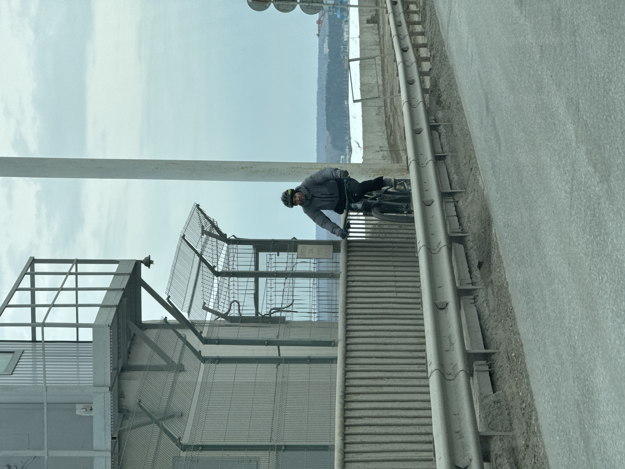 A person on a bicycle is riding on a bridge near an industrial area in Chaikovsky, Russia. The person is wearing a helmet and dark clothing and is leaning slightly forward. The bridge has a metal guardrail and is next to a corrugated metal structure with a cage-like extension on top. A highway is on the right. In the background, a body of water and industrial buildings on the other side are visible under a cloudy sky.