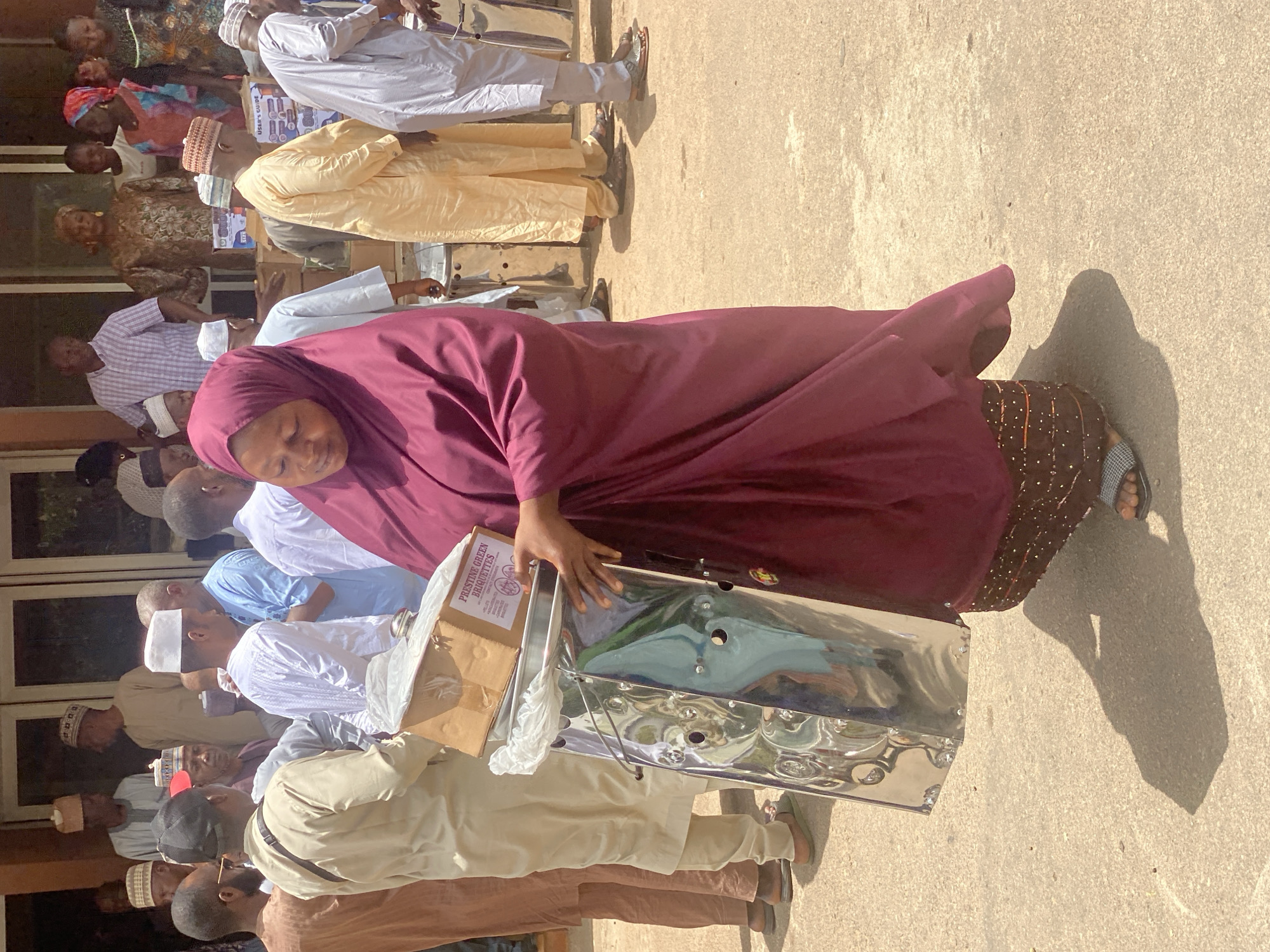 In Unguwar Rimi, Nigeria, a woman wearing a maroon hijab and patterned skirt is carrying a large, silver-colored metal container with attached boxes labeled "Pristine Green Briquettes". She is surrounded by a crowd of men and women, many of whom are wearing traditional Islamic clothing, including white gowns and caps. The location appears to be outdoors on a paved surface.