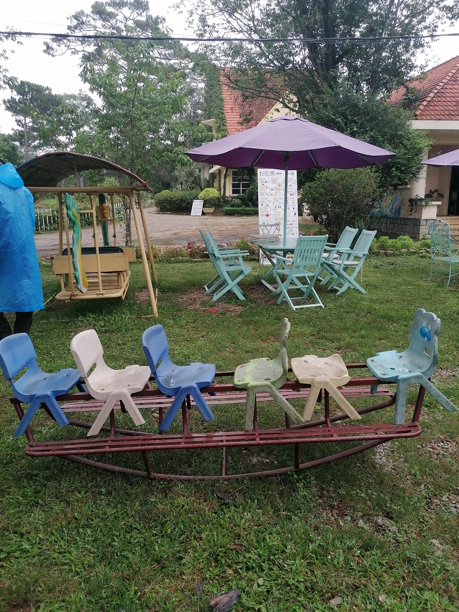 The image displays an outdoor garden area, characteristic of a cafe or restaurant in Da Lat, Vietnam, under an overcast sky. The ground is primarily green grass, appearing damp, suggesting recent rain.
In the foreground, a children's seesaw or rocker is prominent. It features a worn red-brown metal frame supporting six faded plastic chairs in various colors: two blue, one white, one green, one light yellow, and one light blue with a bear-like face.
Towards the left midground, a wooden swing set with a curved canopy is visible. A person partially obscured on the far left wears a bright blue raincoat, indicating cool or wet weather conditions. A white picket fence can be seen beyond the swing set.
On the right midground, several sets of light blue or mint green wooden tables and chairs are arranged on the grass, serving as outdoor dining or seating areas. A large purple umbrella shades one of these tables. Next to the umbrella, an information stand or menu board displays the word "MENU" at its top, though other text is too small to discern. Further back on the right, a single light blue metal chair sits on the grass.
The background is filled with lush greenery, including tall pine trees and other leafy trees, along with a building featuring a distinctive red-tiled roof, common in Da Lat architecture. The scene is static, devoid of active participation, presenting a tranquil, rustic outdoor setting for leisure.