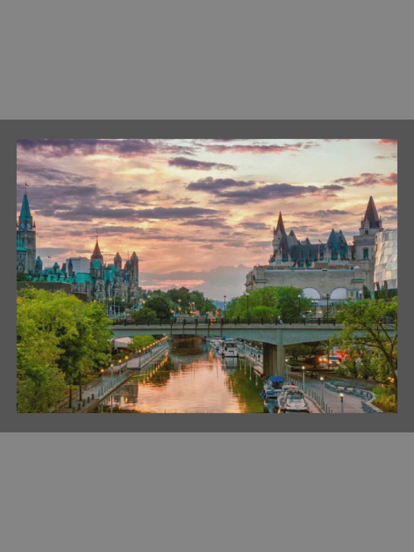 This image presents a picturesque sunset scene over Ottawa, Canada, centered around the historic Rideau Canal. The sky is adorned with soft pink, orange, and purple clouds, indicating twilight or dusk, with hints of the setting sun casting warm light.
On the left side of the frame, the iconic Peace Tower of the Parliament of Canada is visible, along with other stately government buildings featuring teal-colored roofs and gothic architecture, surrounded by abundant green trees. On the right, the grand Fairmont Château Laurier hotel stands prominently, characterized by its distinctive chateau-style architecture and dark spires, with a modern glass-faced building partially visible beside it.
A concrete bridge spans the Rideau Canal, connecting the two sides of the city. Below the bridge, the canal's waters reflect the colorful sky and the surrounding lights. Several recreational boats are moored along the canal's banks, and lampposts illuminate the pedestrian pathways. Small figures of people can be seen walking on the bridge and the canal-side promenades, enjoying the evening. The scene is tranquil and highlights the blending of urban landmarks with natural elements during a beautiful sunset. No visible text is discernable.
