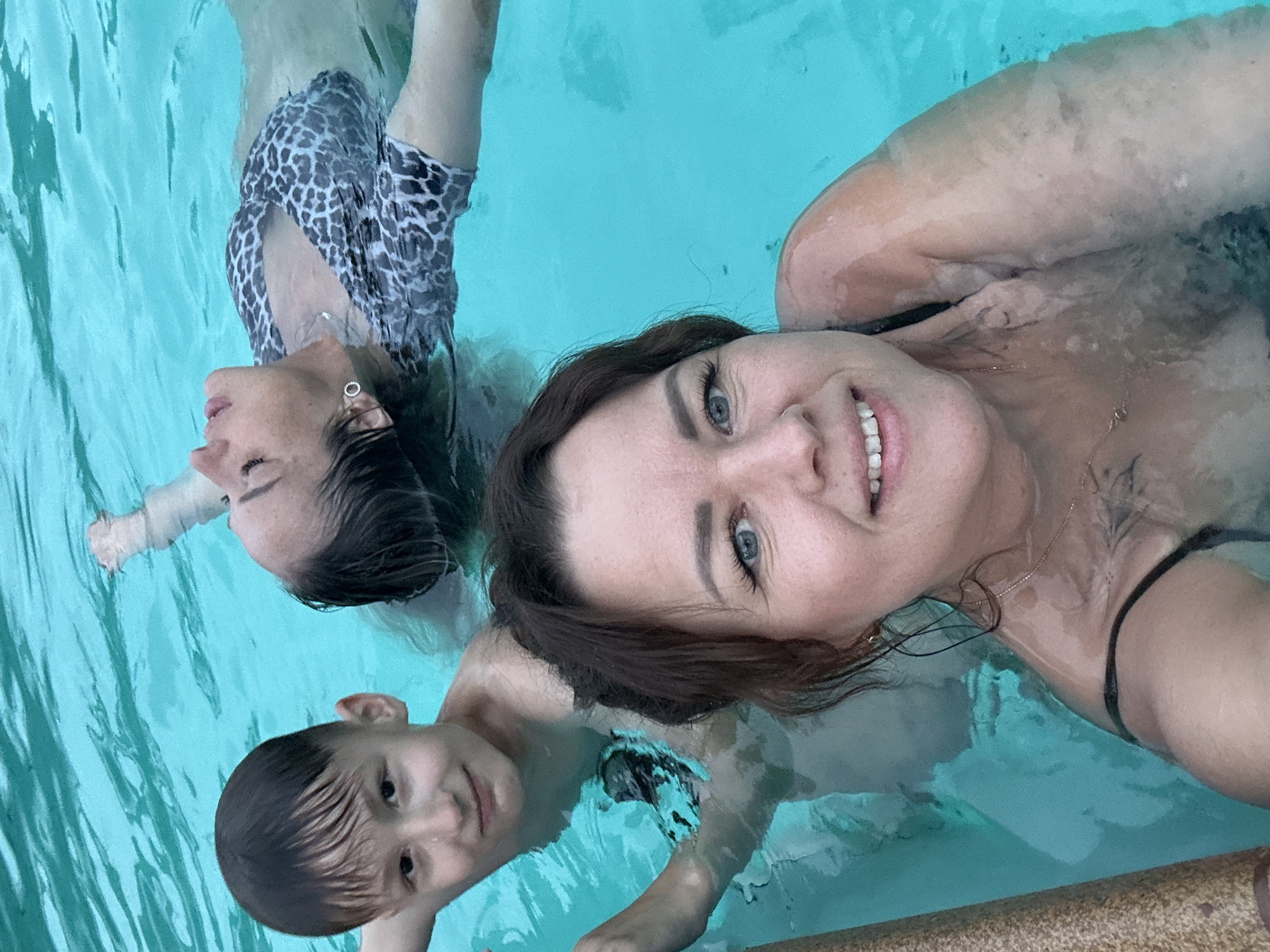 Three individuals are in a swimming pool. A woman in the foreground is smiling at the camera. A second woman in a printed swimsuit is partially submerged behind and to the left of the first. A boy is positioned behind and to the left of the first woman, also partially submerged. The pool water is blue. A pool edge is visible in the lower right corner of the image. There is no indication of the location of Chaikovsky, Russia in the image.