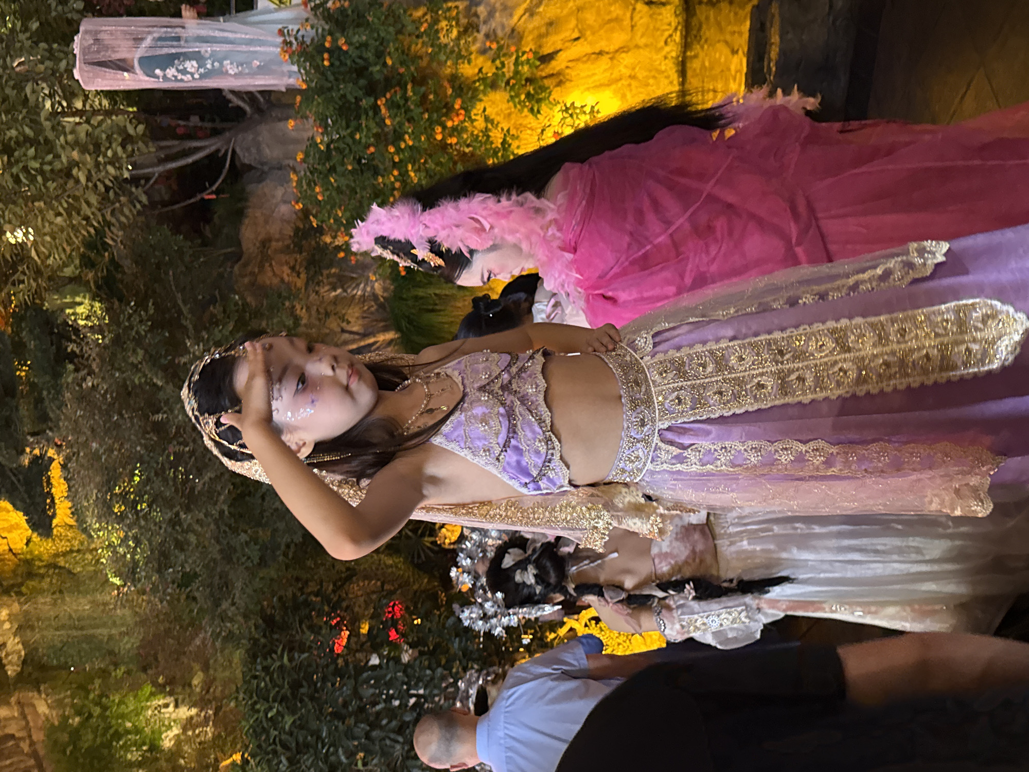 This image shows a group of people dressed in ornate costumes, likely for a performance or cultural event, in what appears to be an outdoor garden setting. In the foreground, a young girl with glitter on her face and elaborate gold and purple attire is posing, with her hand raised to her forehead. To her right, another individual, a woman with long black hair adorned with pink feathers, is wearing a bright pink, flowing garment. Behind them, other individuals in similar festive clothing are visible, along with lush green foliage and a textured, illuminated rock face in the background. A man in a blue shirt is partially visible on the left, observing the scene. The overall impression is of a vibrant, possibly historical or mythological, themed event.