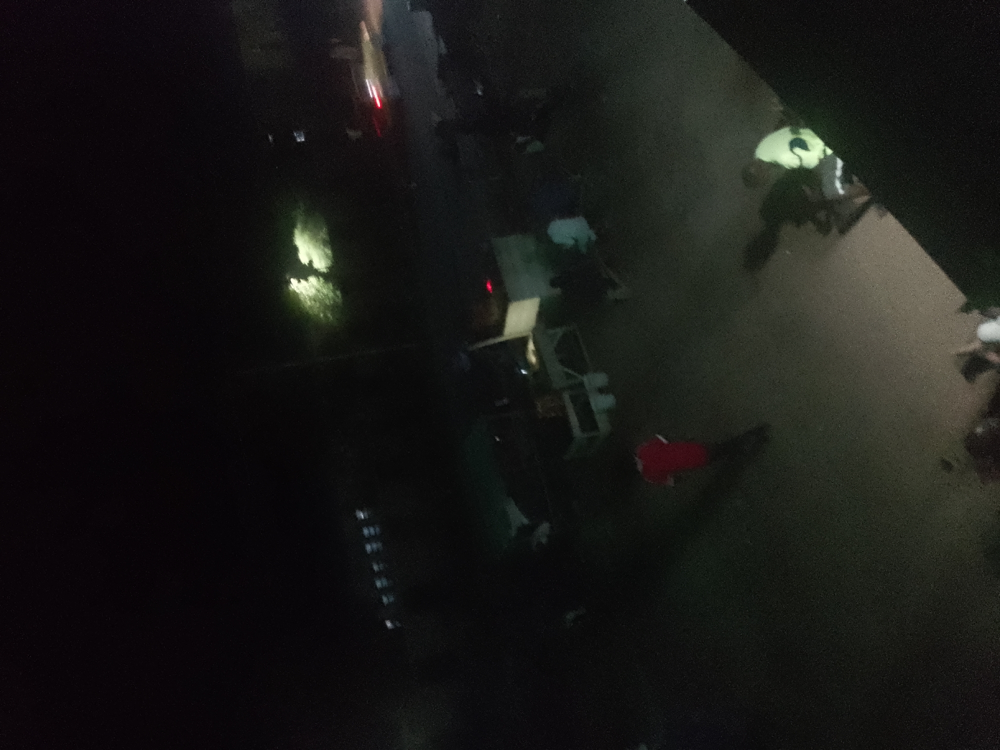 The image captures a nighttime scene in Jalingo, Nigeria, depicting what appears to be a crowded outdoor area with several individuals and various objects. The lighting is dim, with some areas illuminated by what could be artificial lights or reflections, creating a grainy and somewhat blurry visual. In the foreground, a portion of a dark structure or wall is visible. The middle ground features a collection of people, some of whom are wearing bright clothing, including a prominent individual in a neon-green shirt. There are also structures that resemble makeshift stalls or shelters, with one having a white surface and another a red piece of fabric. The ground appears to be wet or reflective, suggesting a possible puddle or flooded area, with scattered lights reflecting on its surface. In the background, there are more indistinct shapes and figures, some of which appear to be in motion. The overall impression is that of a bustling, potentially informal gathering or market scene at night.