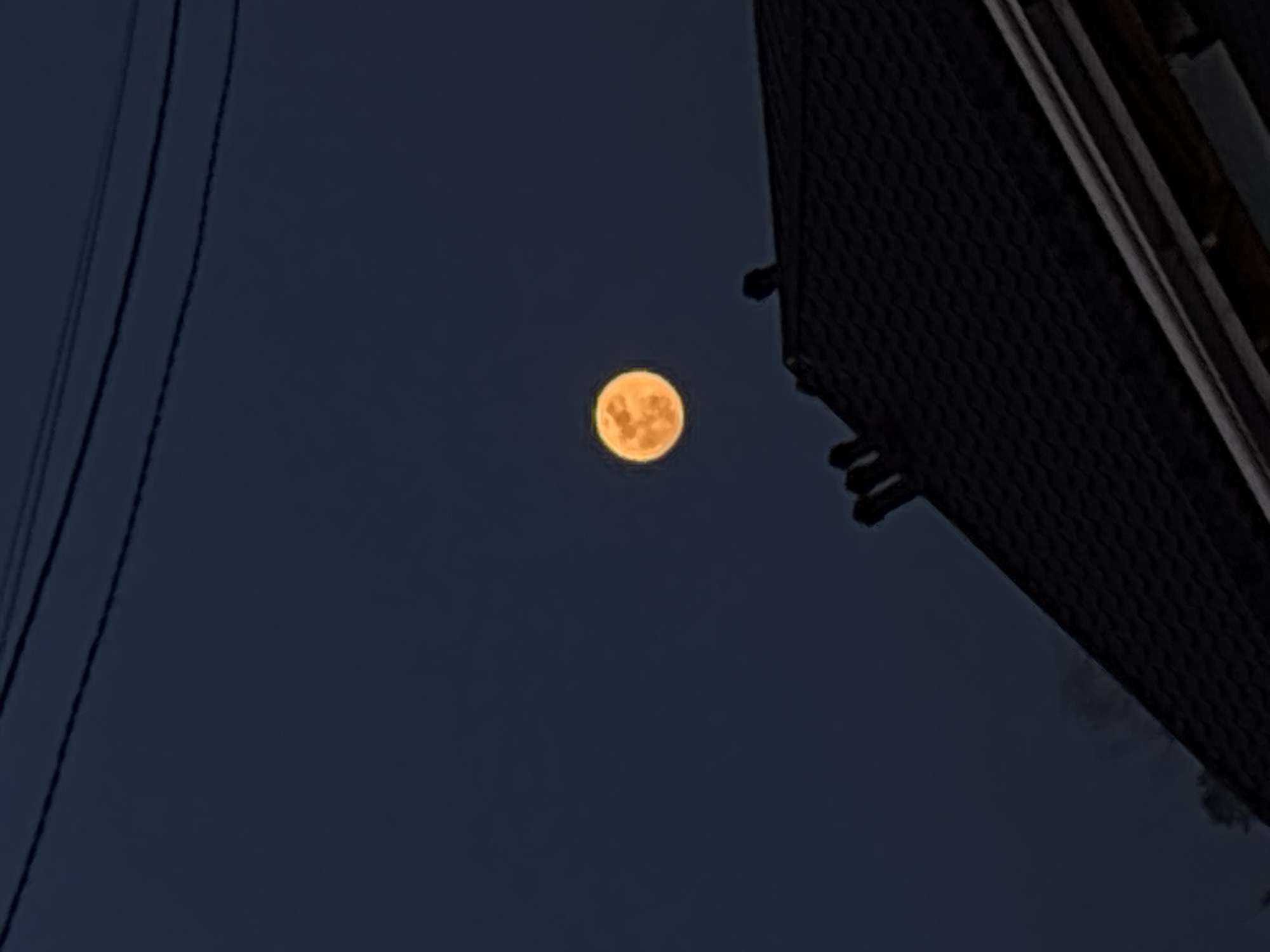 A full moon is centered in the sky, positioned between electrical wires on the left and the corner of a dark building on the right. The sky is a dark blue. The building appears to be several stories tall, and its outer walls have a textured surface.