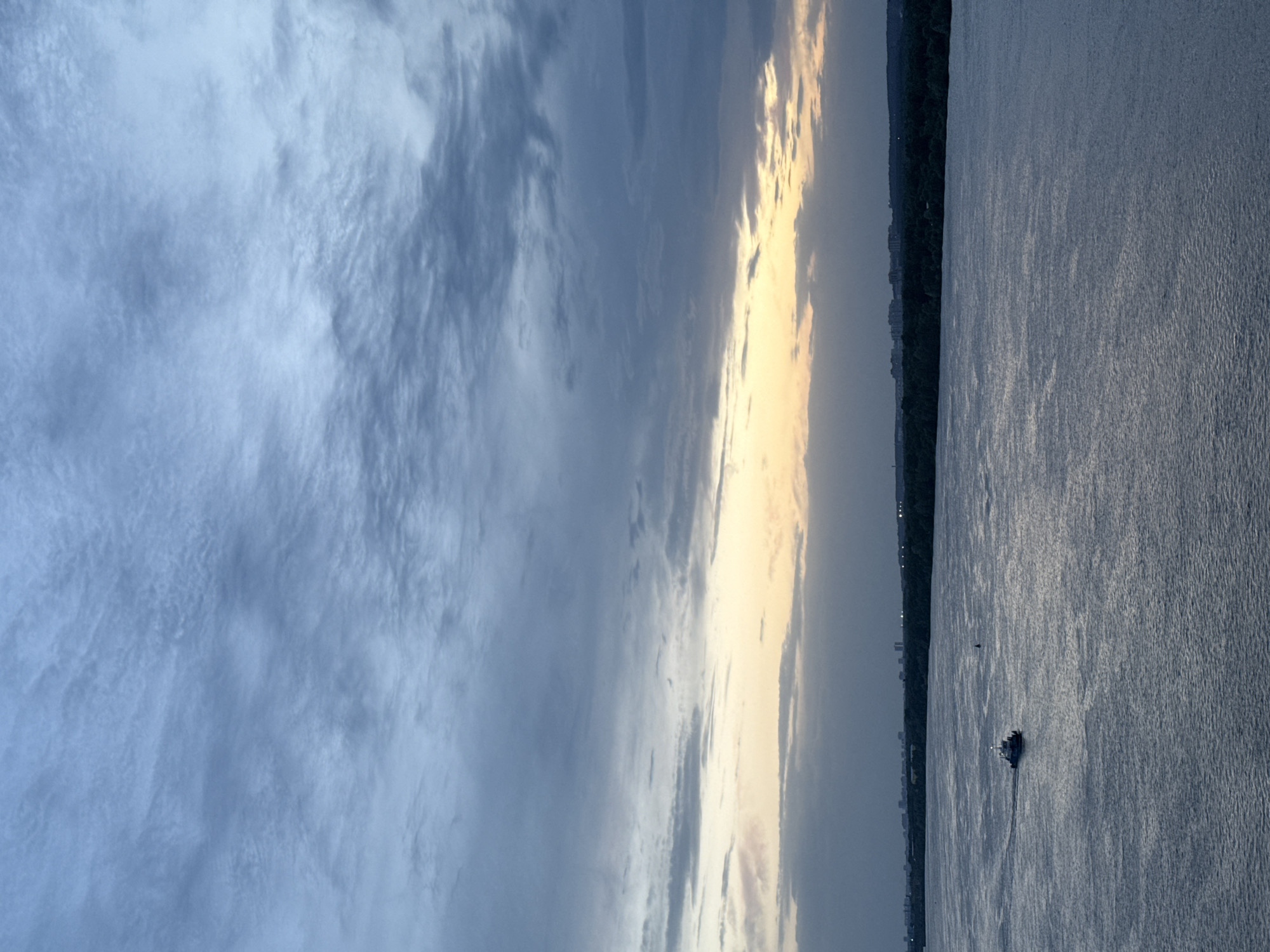 The image shows a body of water, likely the Kama River, in Perm, Russia. The sky is cloudy with a section of bright light, suggesting either sunrise or sunset. A line of trees and buildings can be seen on the horizon. A small boat is visible on the water.