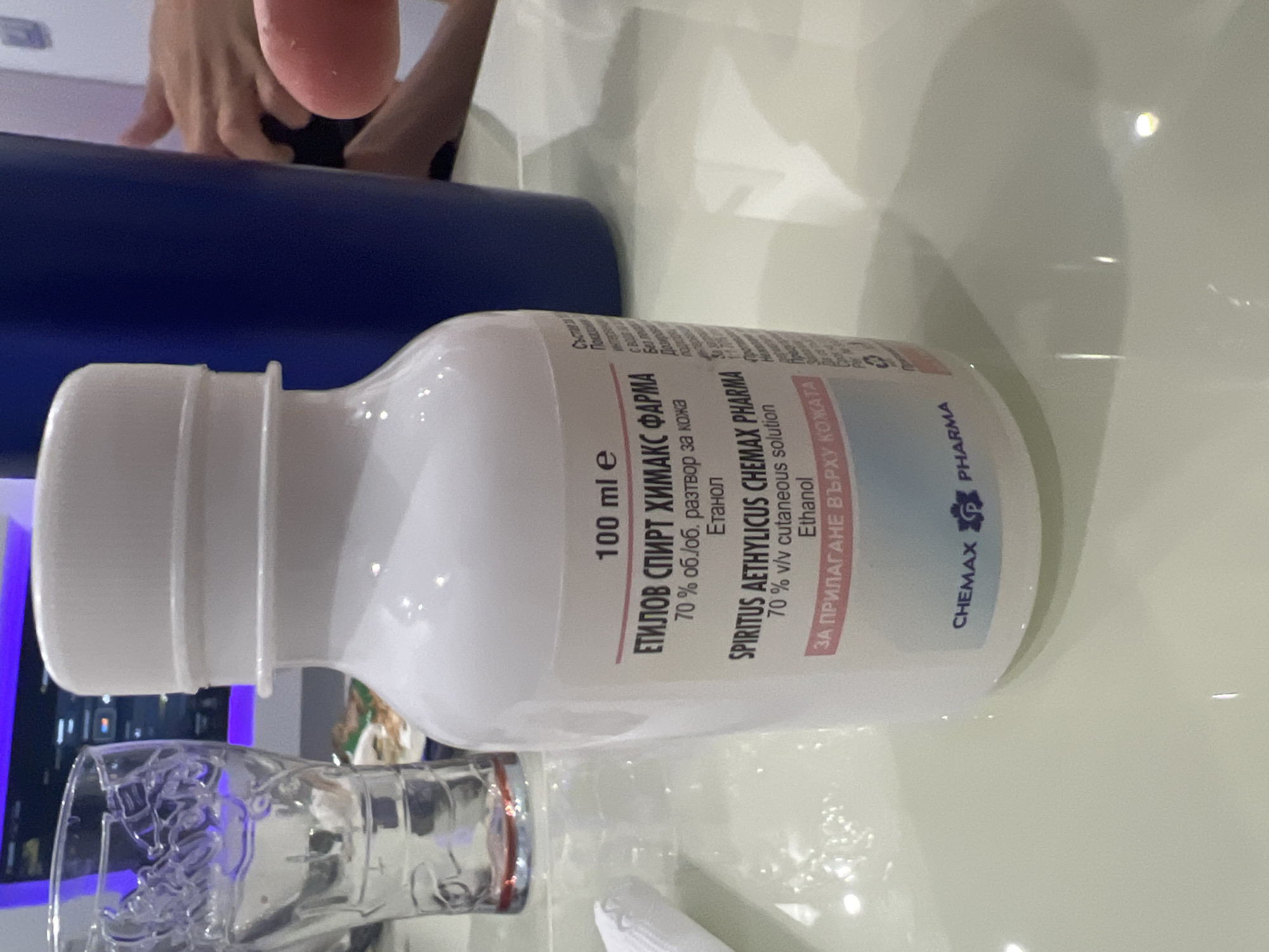 The image shows a bottle of "SPIRITUS AETHYLICUS CHEMAX PHARMA" on a reflective surface. The label indicates it is a 70% v/v cutaneous solution of Ethanol and has a volume of 100 ml. The bottle is white with a white cap. To the left of the bottle is a clear glass with some liquid at the bottom. In the background, a blue cylindrical object is visible, and to its right, two hands are partially visible. The scene appears to be indoors, possibly on a table. The lighting is bright, creating reflections on the surface. The overall setting does not provide specific cues to pinpoint the exact location within Podgumer, Bulgaria, other than being an indoor environment with common household or personal items.
