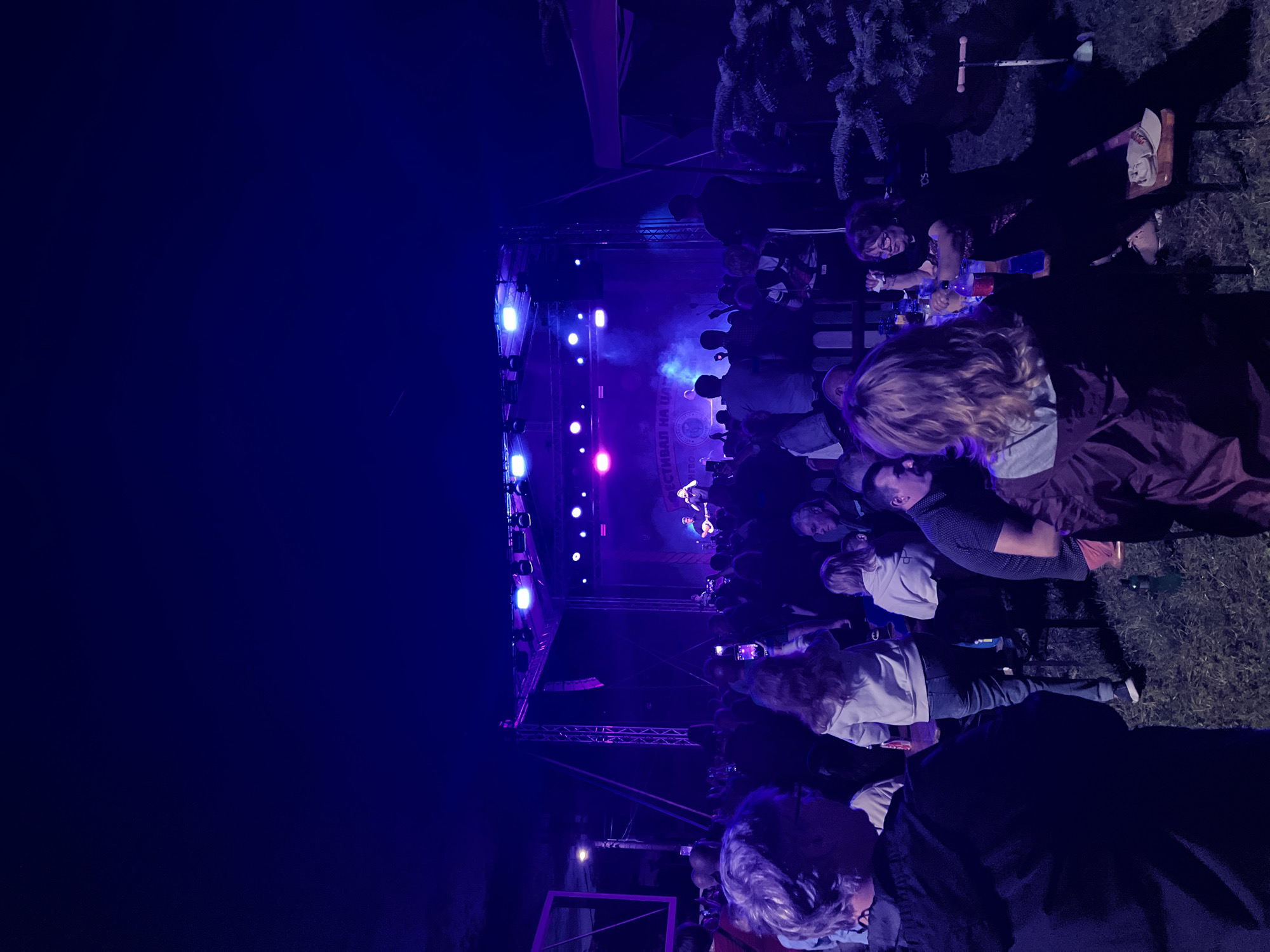 This image depicts an outdoor night event, likely a festival or concert, in Kranevo, Bulgaria. A raised stage dominates the midground, illuminated by multiple purple and blue theatrical lights affixed to a metal truss structure. Two performers are visible on stage, one holding a guitar, against a backdrop displaying Cyrillic text, including "ФЕСТИВАЛ НА" (Festival of). Atmospheric haze or smoke is present around the stage area, highlighted by the lighting.
A large crowd stands directly in front of the stage, with many individuals holding up smartphones, presumably recording the performance. A large, rectangular, purple-lit structure is partially visible on the far left. A low wooden fence separates the standing crowd from a foreground area with seated individuals.
In the immediate foreground, a grassy terrain is visible. On the far left, a person with short, light-colored hair and glasses, wearing a dark hooded jacket, is seen from the back-left shoulder, facing towards the right. Towards the center, a person with long, light-brown hair, wearing a light-colored jacket and blue jeans, stands or walks away from the viewer. A man is seated to their right. On the right side, a group of people are seated at a low table or bench. A woman with red hair and glasses, wearing a dark shirt, smiles while seated at the table, which holds multiple bottles and cups. To her left, another person with light-colored hair, wearing a dark jacket, is seen from behind. A dark evergreen bush is present on the far right. A green bottle lies on the grass in the center-right foreground, and a small metal kick scooter is on its side in the bottom right corner.