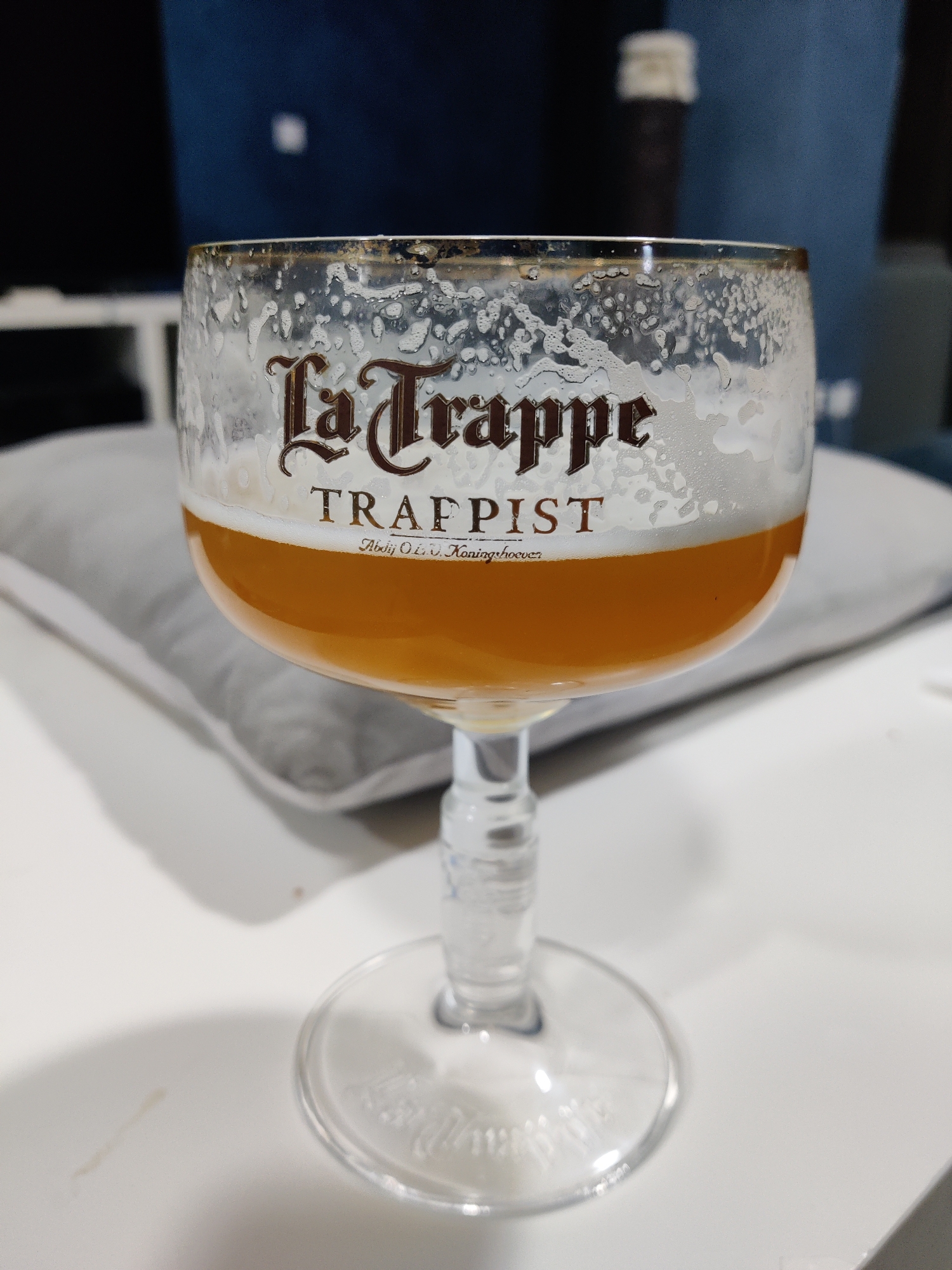A glass of La Trappe Trappist beer sits on a white table. The beer is a golden color with a foamy head, and the glass is decorated with the La Trappe logo. The location appears to be indoors, possibly a residence, as evidenced by the presence of a gray pillow and a blurred background that suggests furniture and other household items. The city and specific location are unknown.