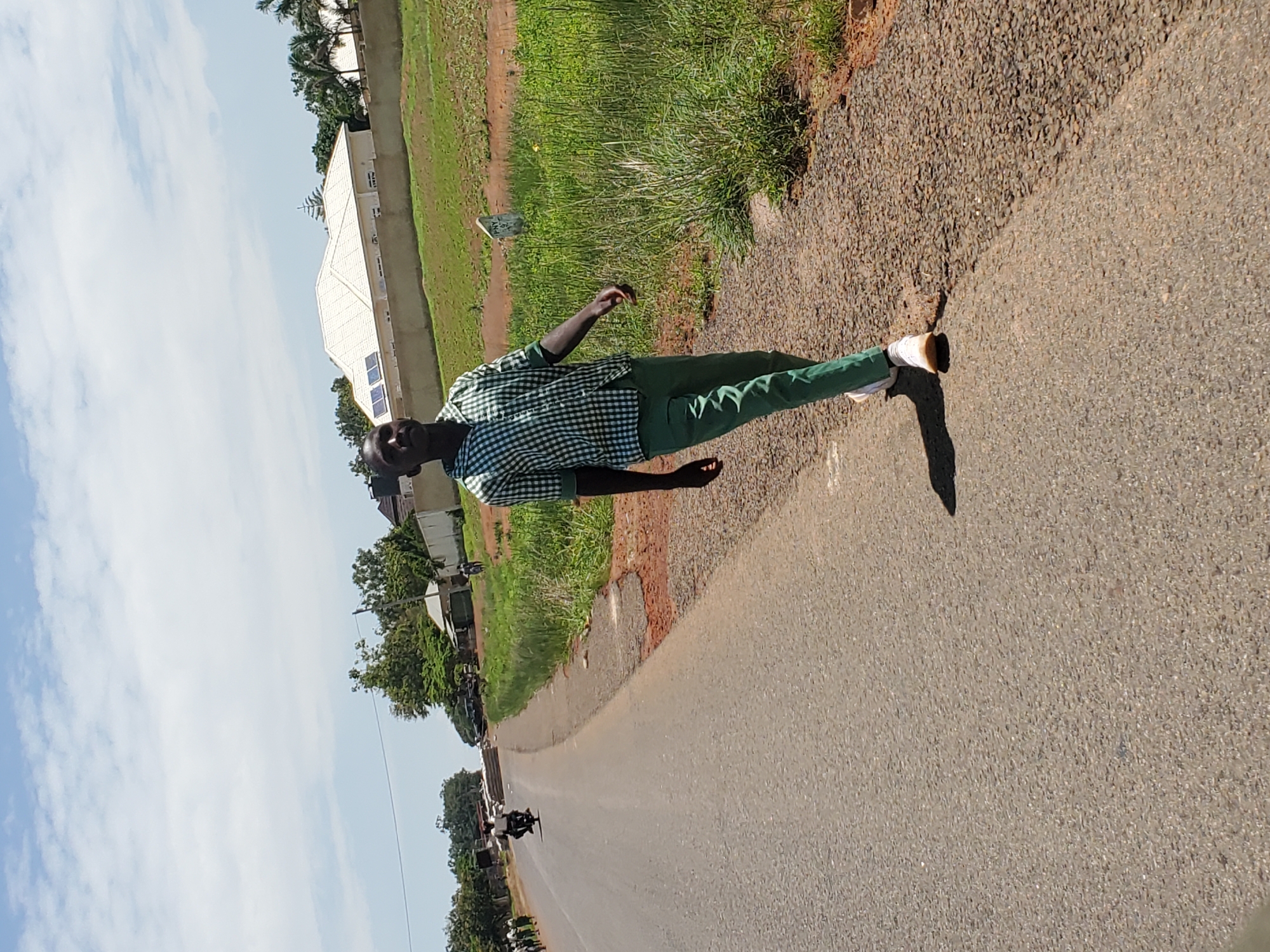 A young Black male, wearing a green and white checkered shirt and green pants with white sneakers, walks along the shoulder of a paved road in Pankshin, Nigeria. He is facing towards the right of the frame and looking upwards and to the right. The road is asphalt, with a gravel shoulder on the right side of the image. Beyond the shoulder is a grassy area with some dry patches and a small white marker post. In the background, a modern house with a white roof and solar panels is visible, surrounded by a low concrete wall. Palm trees and other vegetation are scattered in the background, and a bright blue sky with scattered white clouds dominates the upper portion of the image. Other people and a motorcycle are visible further down the road, suggesting a populated area. The lighting indicates it is daytime, likely mid-morning or afternoon.