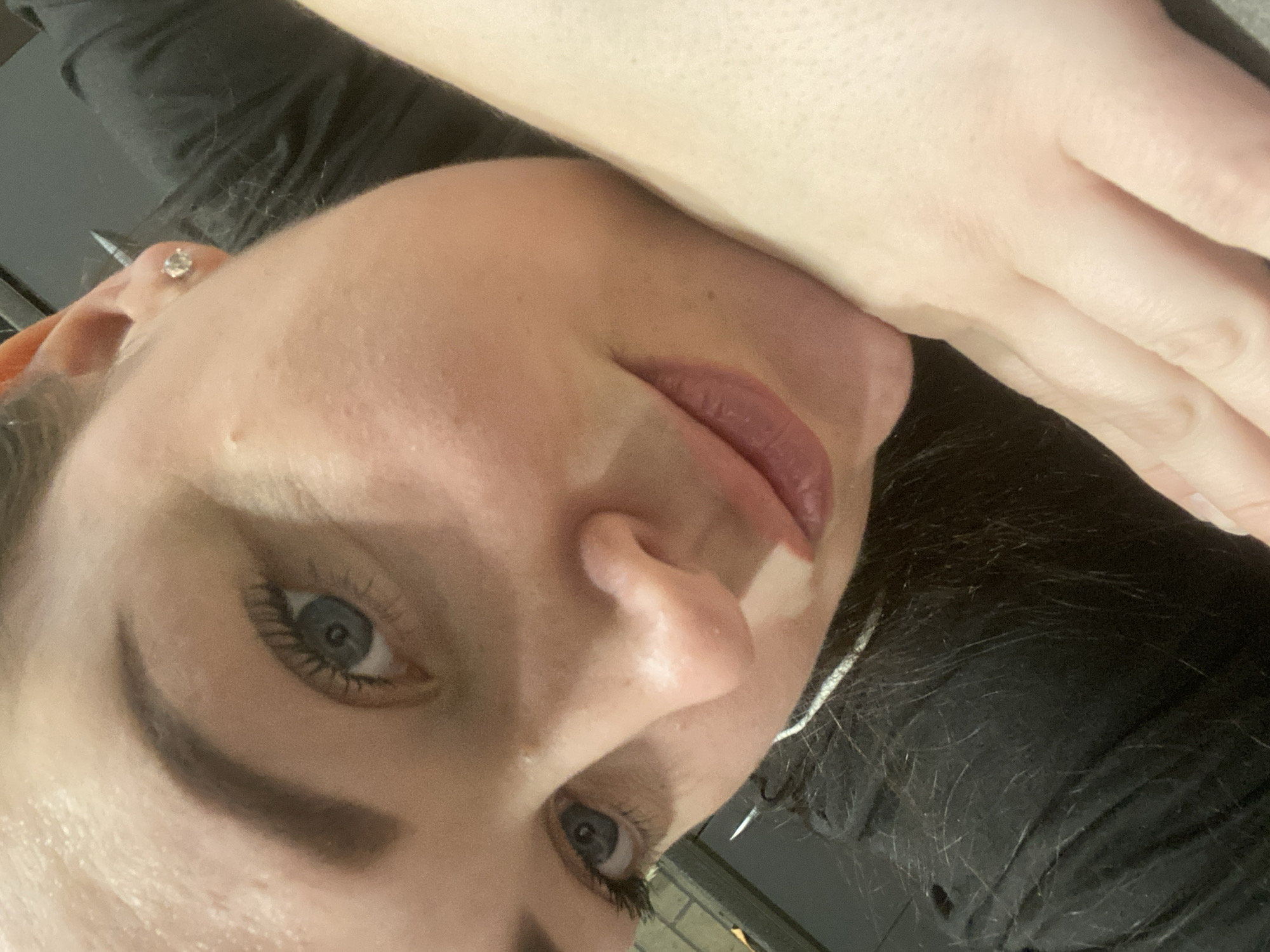 A close-up portrait of a woman with fair skin, blue eyes, and dark eyebrows and eyelashes. She has a silver stud earring in her ear. Her lips are painted with a pink lipstick. She is wearing a black garment, and her hand is visible. The location cannot be determined from the image.