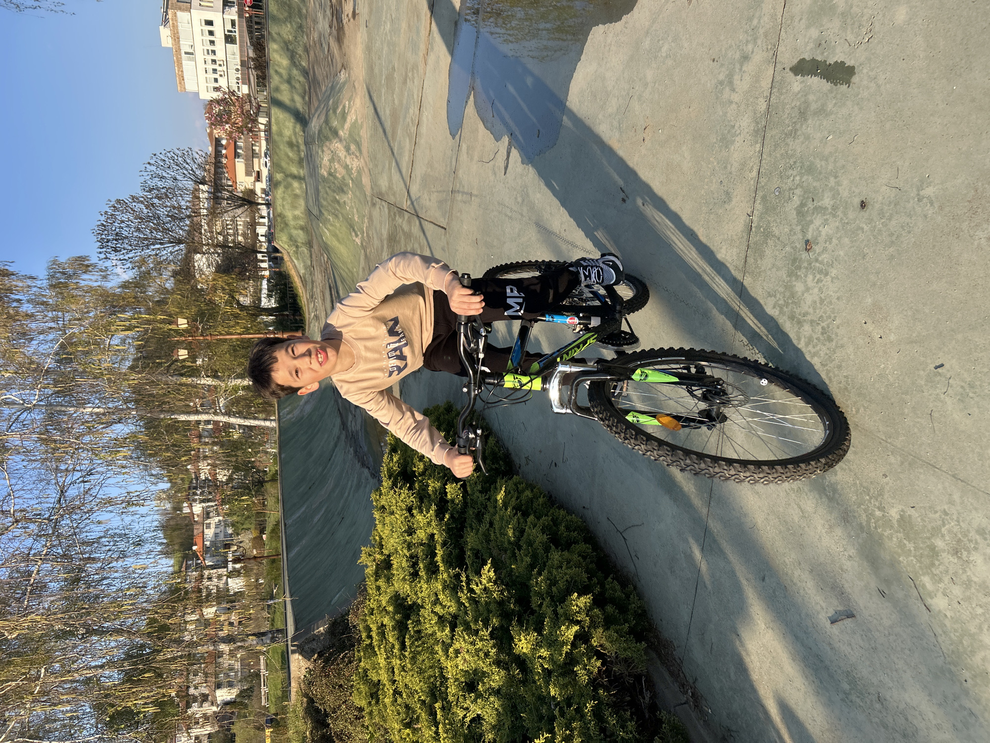 A young boy is standing on a bicycle in an outdoor, urban environment. The bicycle is black and green. The boy is wearing a beige shirt with black pants and black and white shoes. He appears to be smiling. The ground is paved with concrete. Behind the boy is a green shrub and trees with bare branches. In the background, buildings are visible. This scene is plausibly located in Zlatograd, Bulgaria.