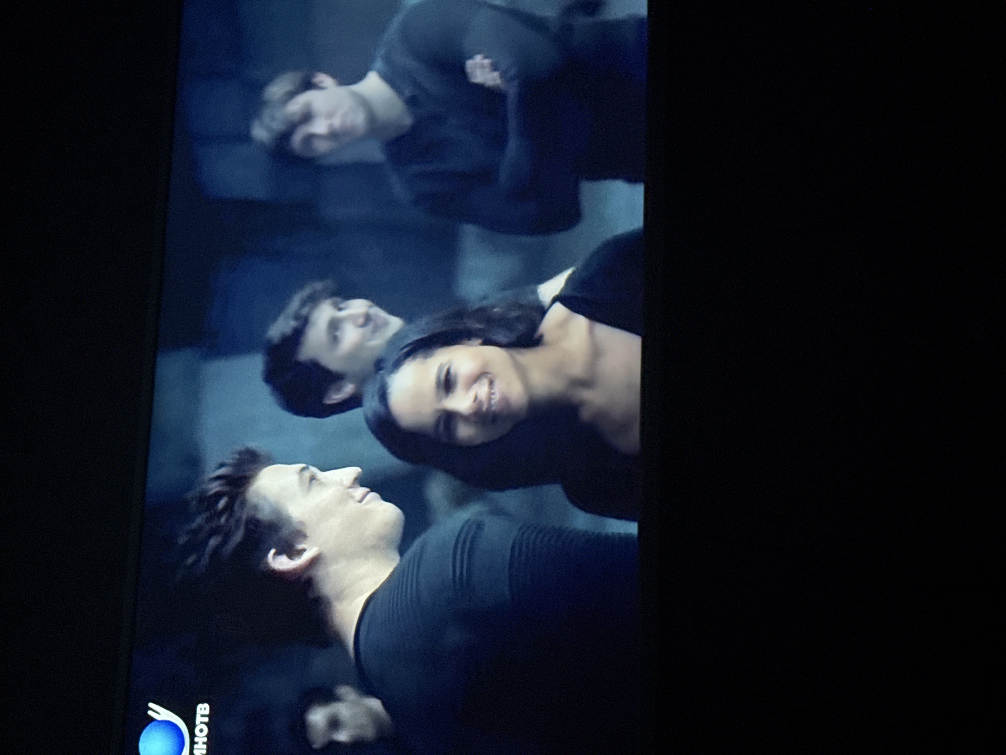 The image shows a screen displaying a scene with four people. Three are in the foreground: a woman with dark hair is smiling, and two men are facing away from her. A fourth man is visible in the background. The setting appears to be a dark, possibly indoor, environment. A television channel logo is visible in the lower left corner, with the text "ИНОТВ" adjacent to it. The image does not contain information about Mytishchi, Russia.
