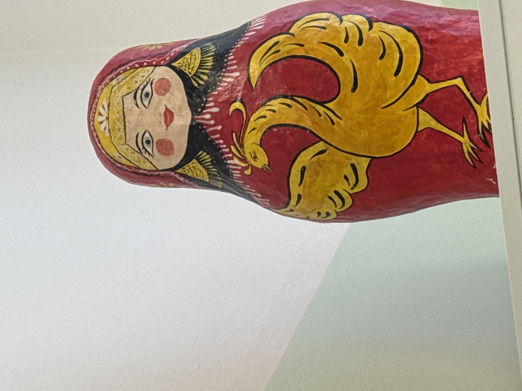 A photograph shows a painted, wooden Russian nesting doll, also known as a matryoshka doll. The doll features a woman's face painted on the front, with a yellow headscarf and red cheeks. The doll is red, with a yellow image of a mythical bird on the side. The doll is placed against a white and light green background. The location of the photograph is unknown.