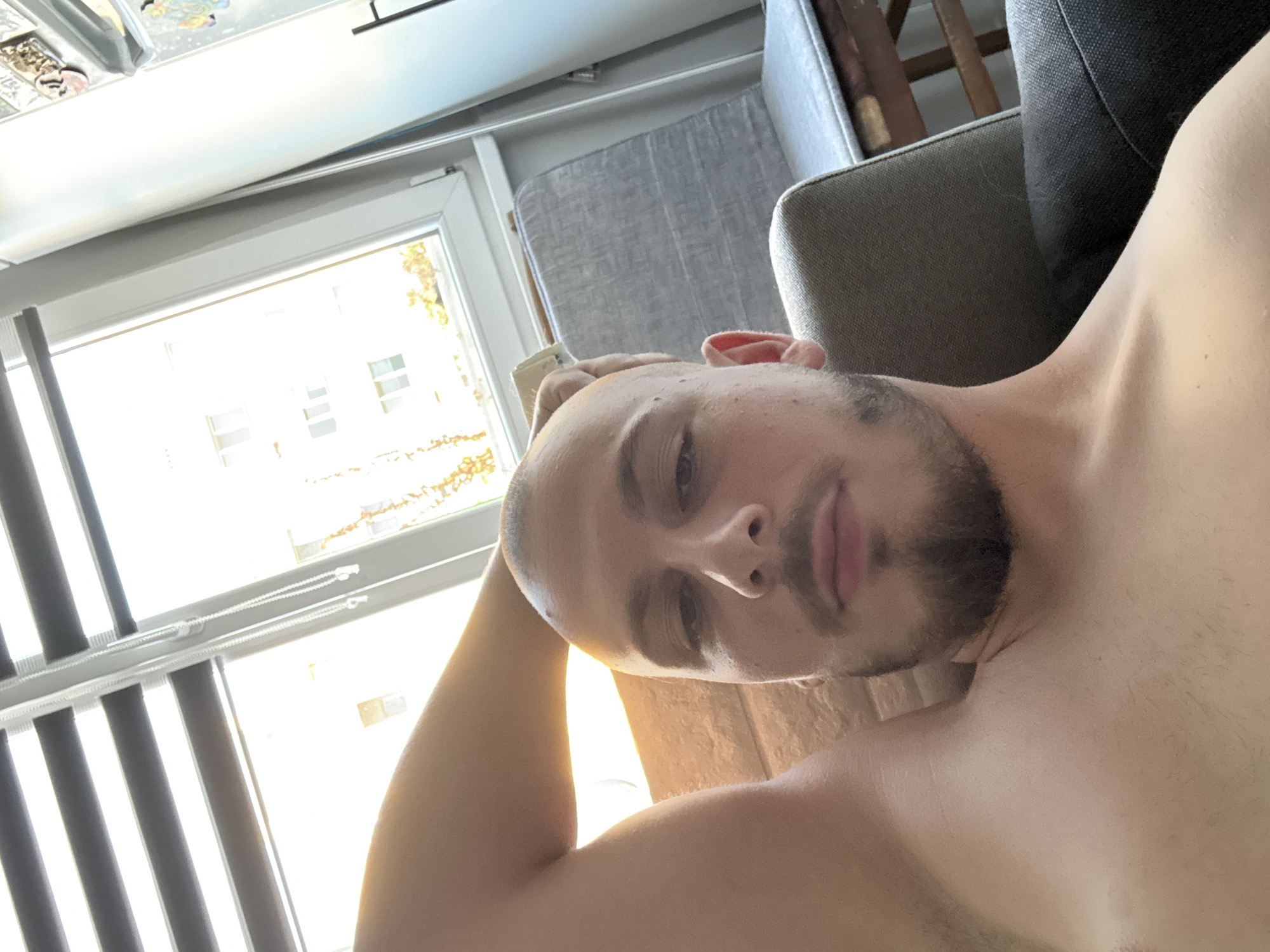 A young, bald man with a goatee is lying on a sofa indoors, with his left arm behind his head. Sunlight streams in through a window, illuminating the room. The man is shirtless. A grey armchair is visible to his right, and a wooden stool is partially visible behind it. Outside the window, a brick building is visible, along with trees and a blue sky. The overall lighting suggests it is daytime.