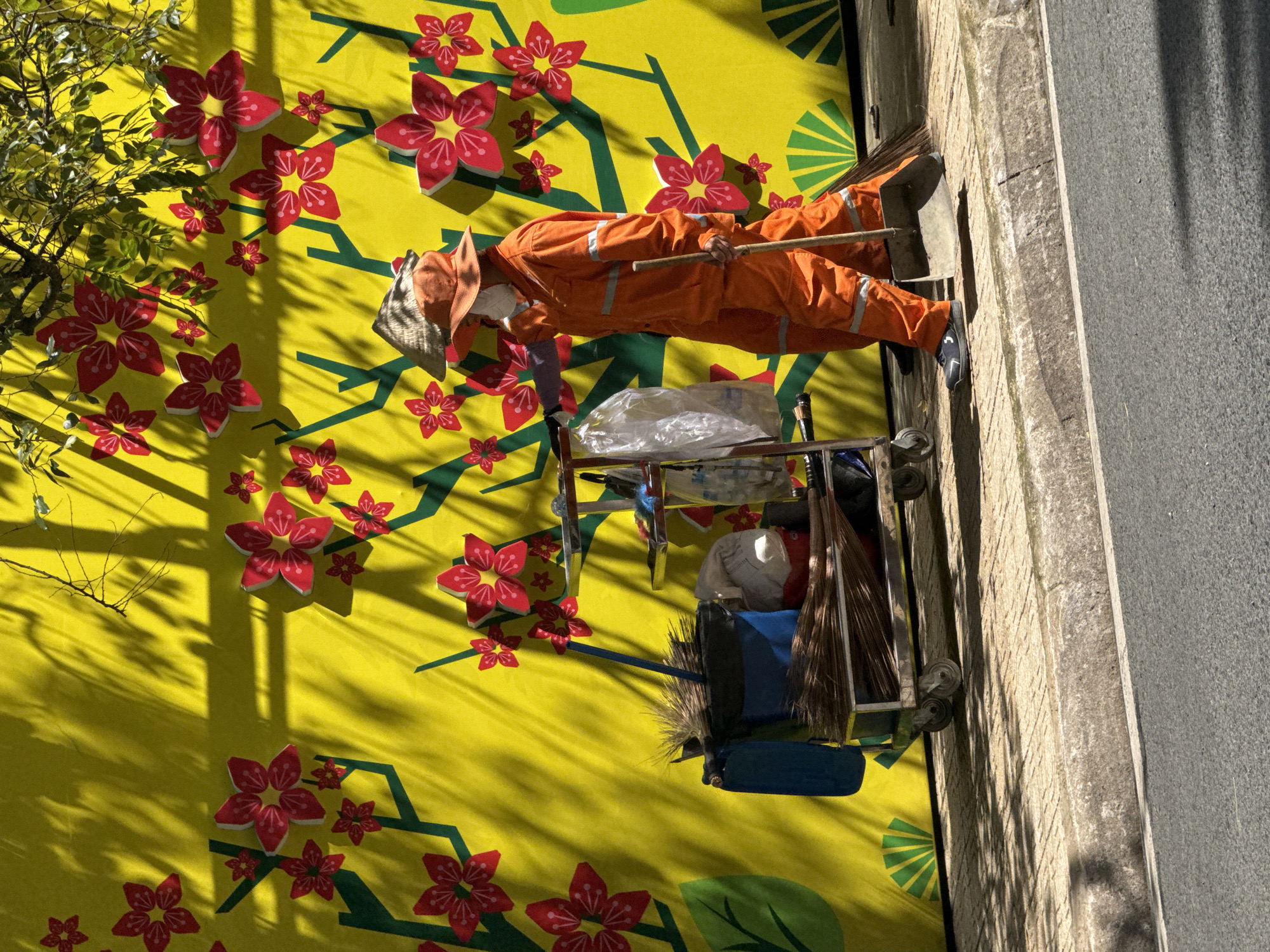 The image depicts a street scene in Da Lat, Vietnam, with a prominent yellow mural featuring red floral motifs and green branches. In the foreground, a street cleaner, dressed in an orange uniform and wearing a conical hat and a face mask, pushes a cart filled with brooms and cleaning supplies. The cart has a blue bag and a plastic bag. The cleaner is holding a broom and a dustpan, and appears to be in motion, walking along the sidewalk. The sidewalk is paved with textured tiles and bordered by a curb. Shadows from trees are cast across the mural and the sidewalk, indicating a sunny day. The street in the lower portion of the image is asphalt.