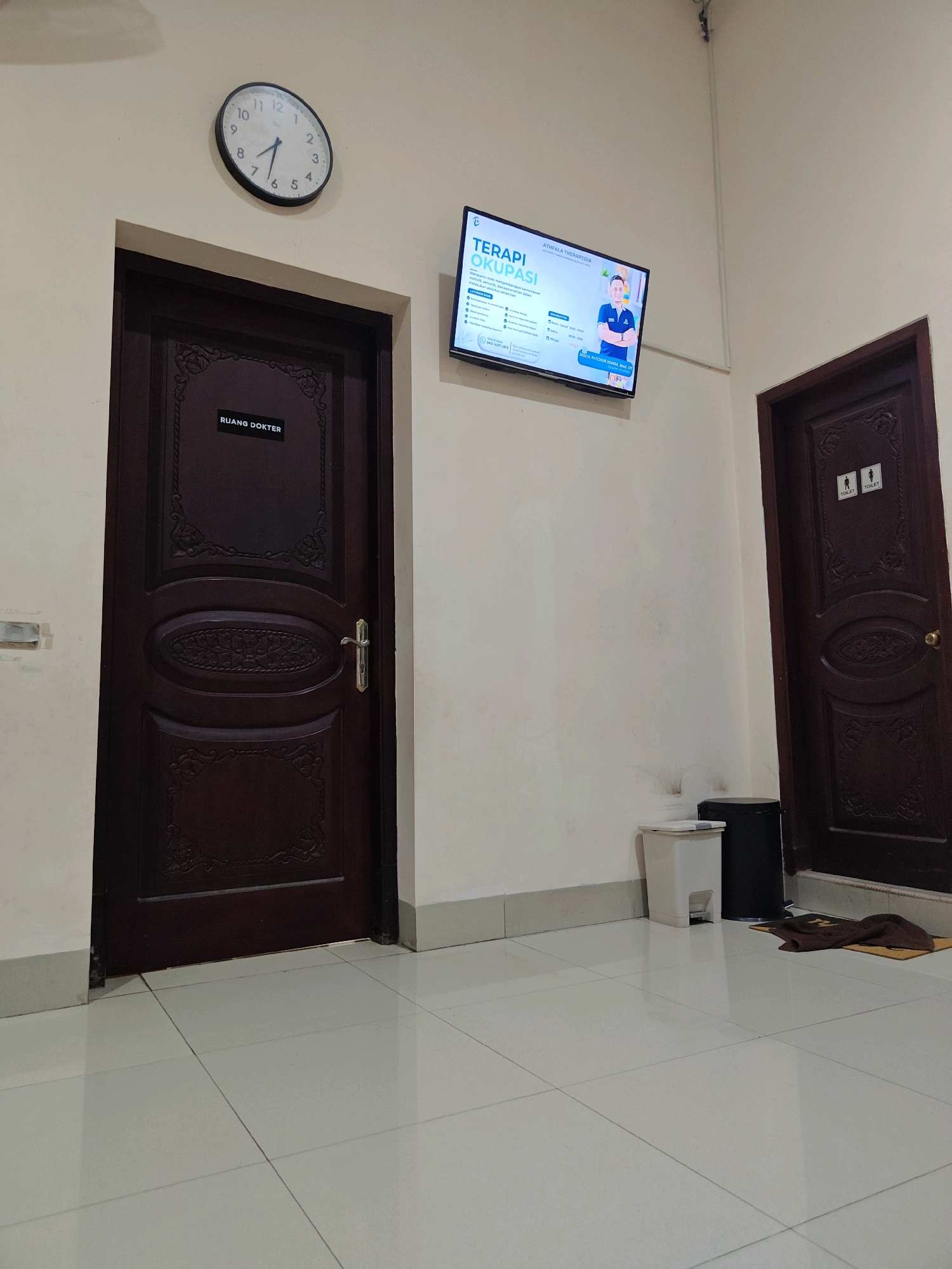 The image shows a hallway with two doors. The door on the left has a sign that reads "RUANG DOKTER," which translates to "Doctor's Room." The door on the right has "TOILET" signs with male and female figures above them. A television screen is mounted on the wall between the two doors, displaying information about "TERAPI OKUPASI" (Occupational Therapy). A round clock is visible on the wall above the doctor's room door. Two trash cans, one white and one black, are placed near the toilet door. The floor is tiled with light-colored square tiles. The scene appears to be inside a clinic or medical facility. While the image does not explicitly state its location in Situbondo, Indonesia, the use of Indonesian language on the signs suggests it is in Indonesia.