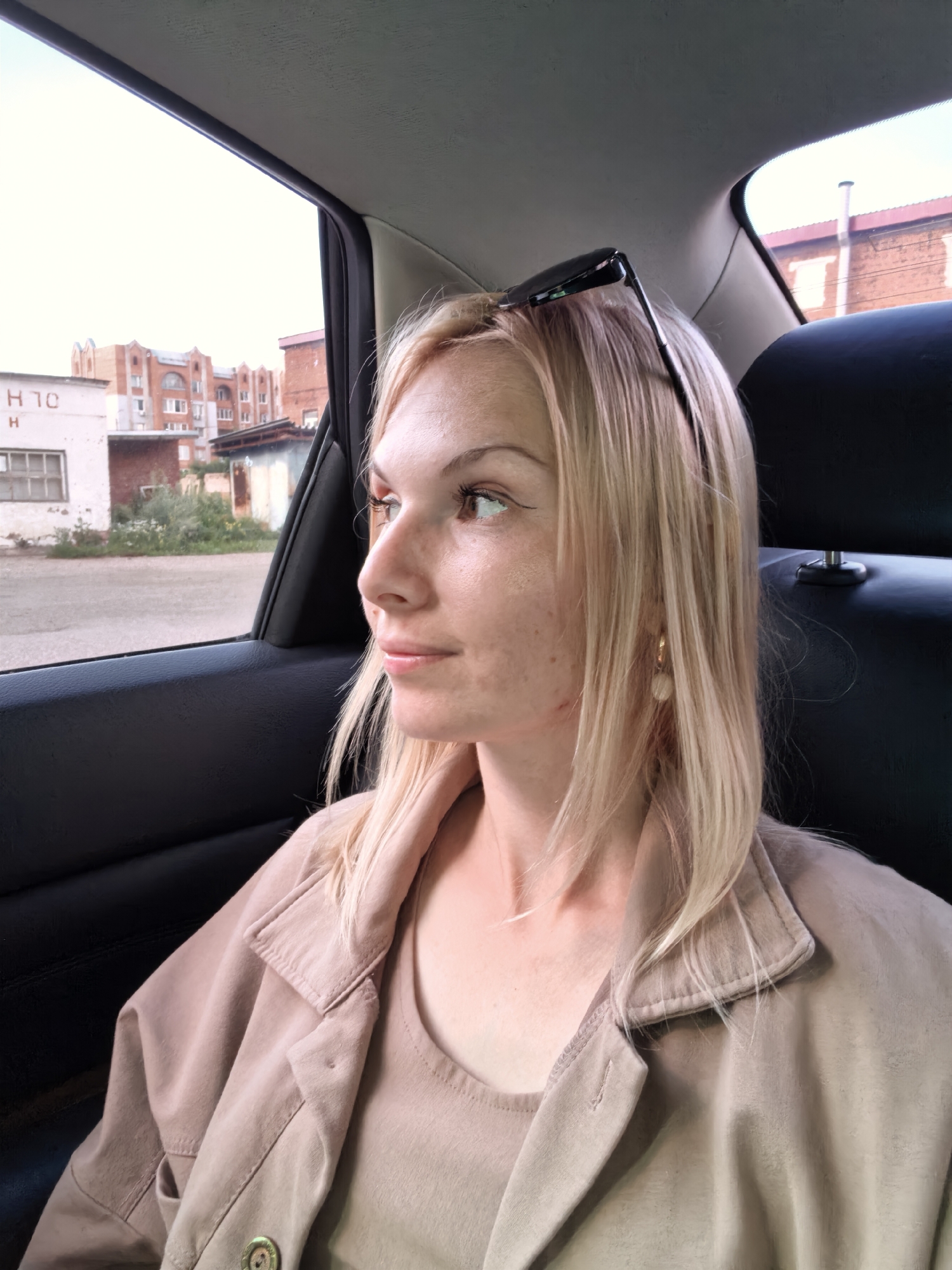 A young blonde woman with fair skin and freckles is seated in the backseat of a car, looking out the window. She has her sunglasses perched on top of her head. She is wearing a light brown jacket over a matching shirt and a gold hoop earring with a pearl dangle. The car interior is visible, with dark seats and part of the ceiling. Outside the window, industrial buildings and a dirt road are visible, with a brick building in the background. The image appears to be taken in an urban or industrial area, possibly on the outskirts of Sterlitamak, Russia.