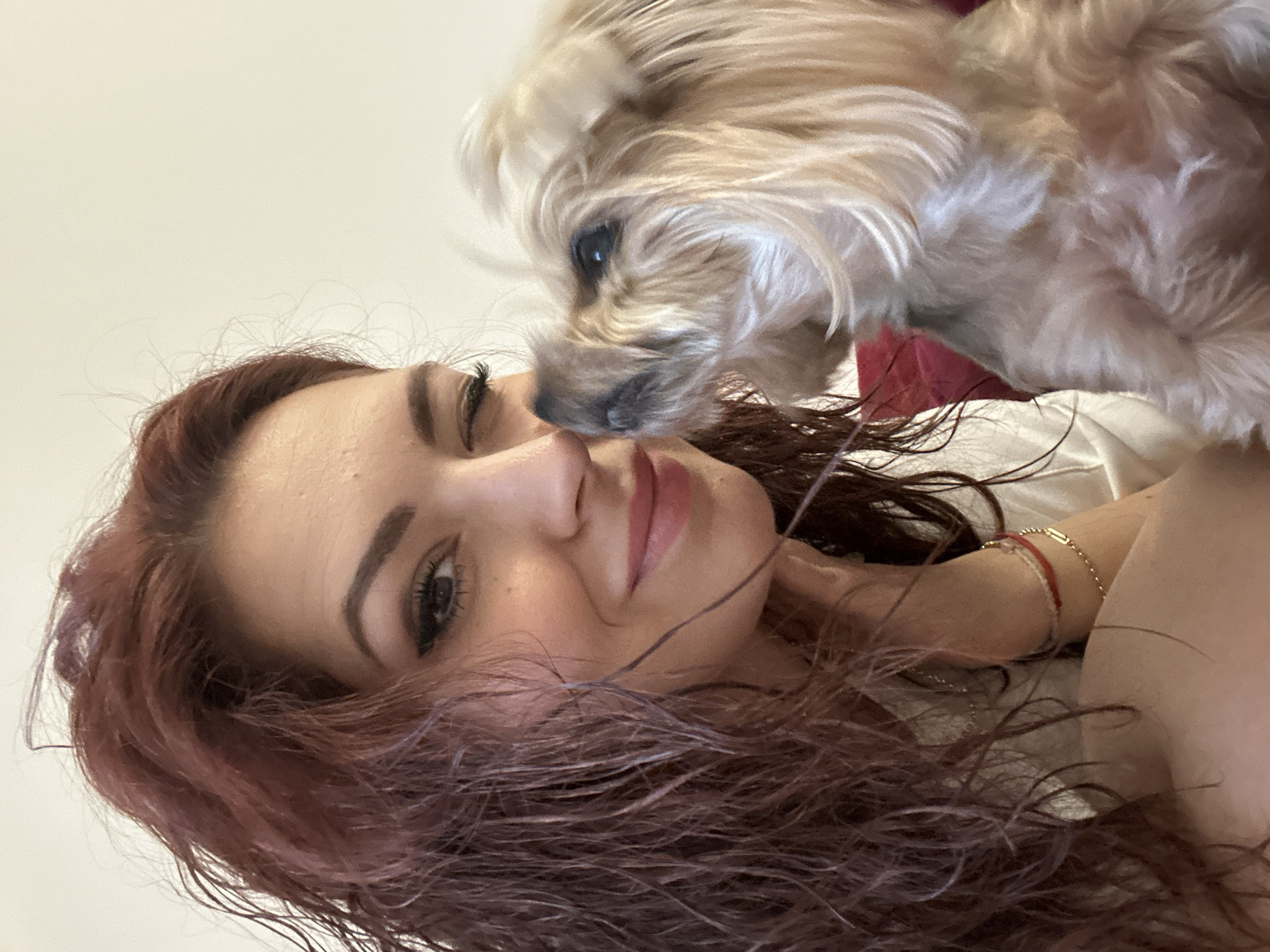 An indoor close-up selfie, captured in Sofia, Bulgaria, features a woman with long, wavy, reddish-brown hair and a small, light-tan colored dog. The woman, positioned on the left side of the frame, is smiling with her left eye open and her right eye in a playful wink. She has dark eye makeup and light rose-colored lipstick. Her left wrist, visible near the bottom center, wears a red string bracelet and a delicate gold-colored chain bracelet. The small dog, likely a Yorkshire Terrier with long, wispy fur, is on the right side, nuzzling its dark nose against the woman's cheek and looking upwards towards her. A plain, light beige wall forms the background, with a small section of red fabric partially visible behind the dog's back and to the left of the woman's shoulder.